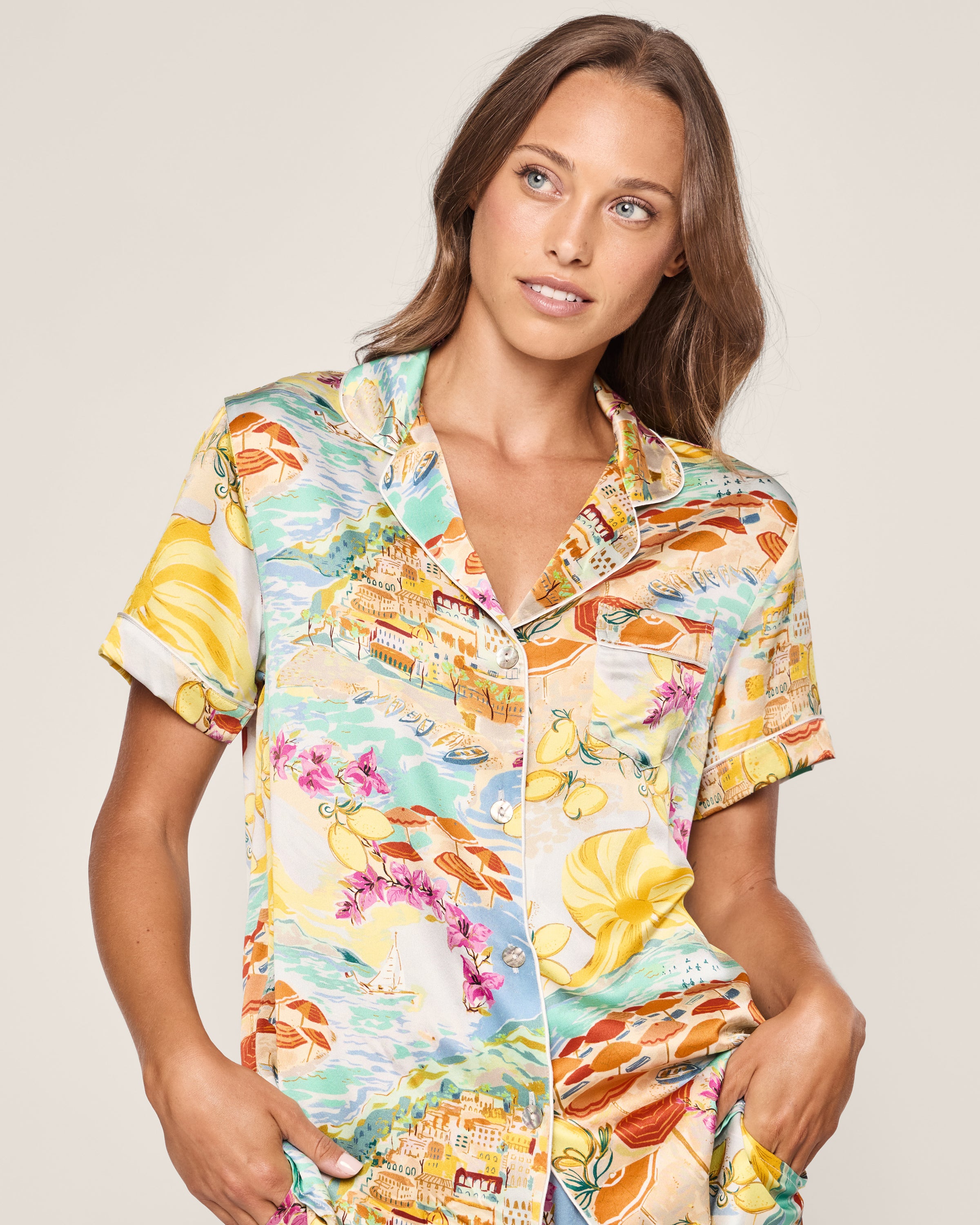 A woman in Petite Plumes Womens Silk Short Set in La Dolce Vita, featuring vibrant beach motifs like lemons, bicycles, flowers, and umbrellas on a light background, gazes aside with a relaxed expression.