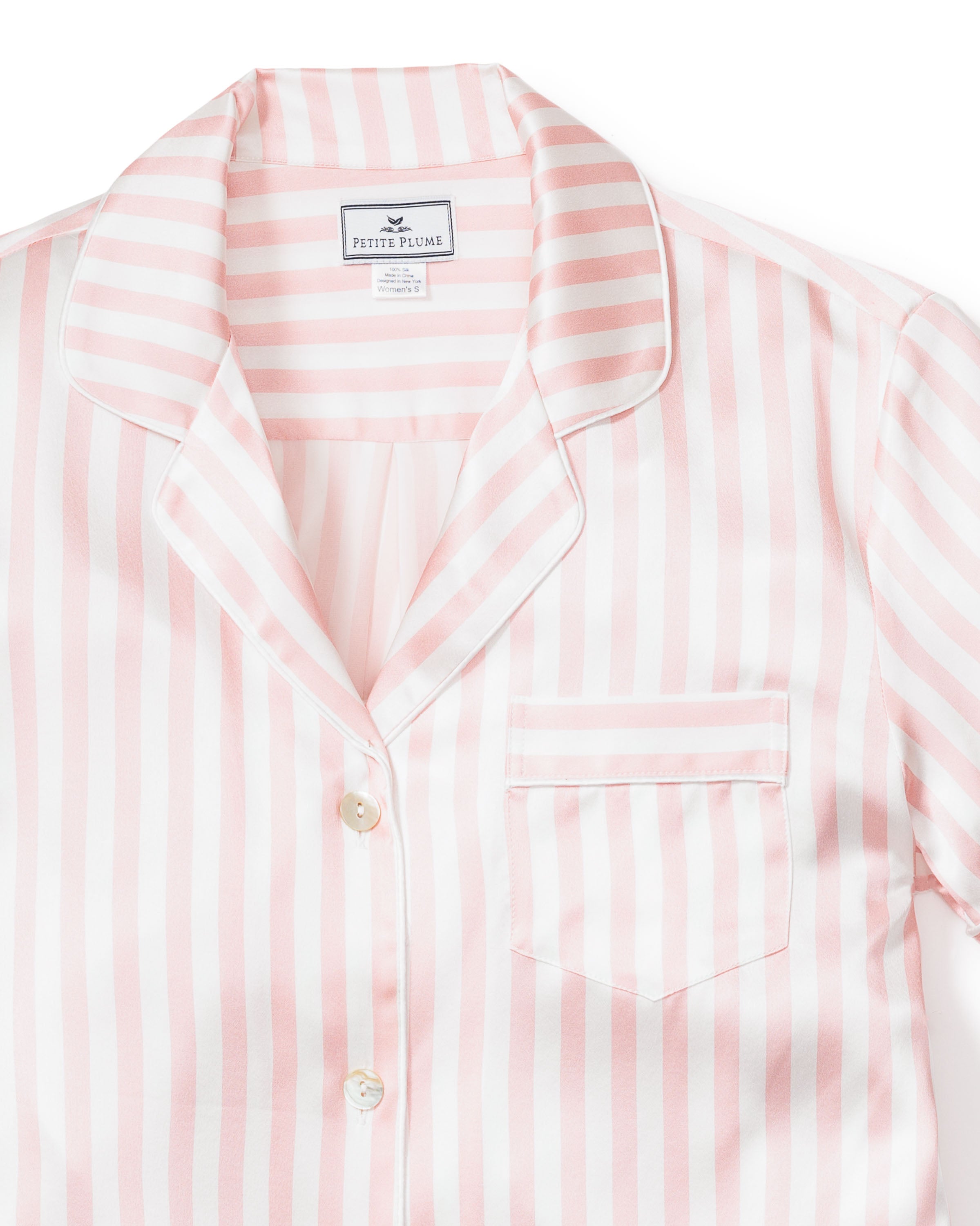 A close-up shows the Womens Silk Pajama Short Set in Pink Stripe by Petite Plume, featuring a collared top with a chest pocket and pearl-colored buttons. Made from Mulberry silk, the fabric looks silky and luxurious.