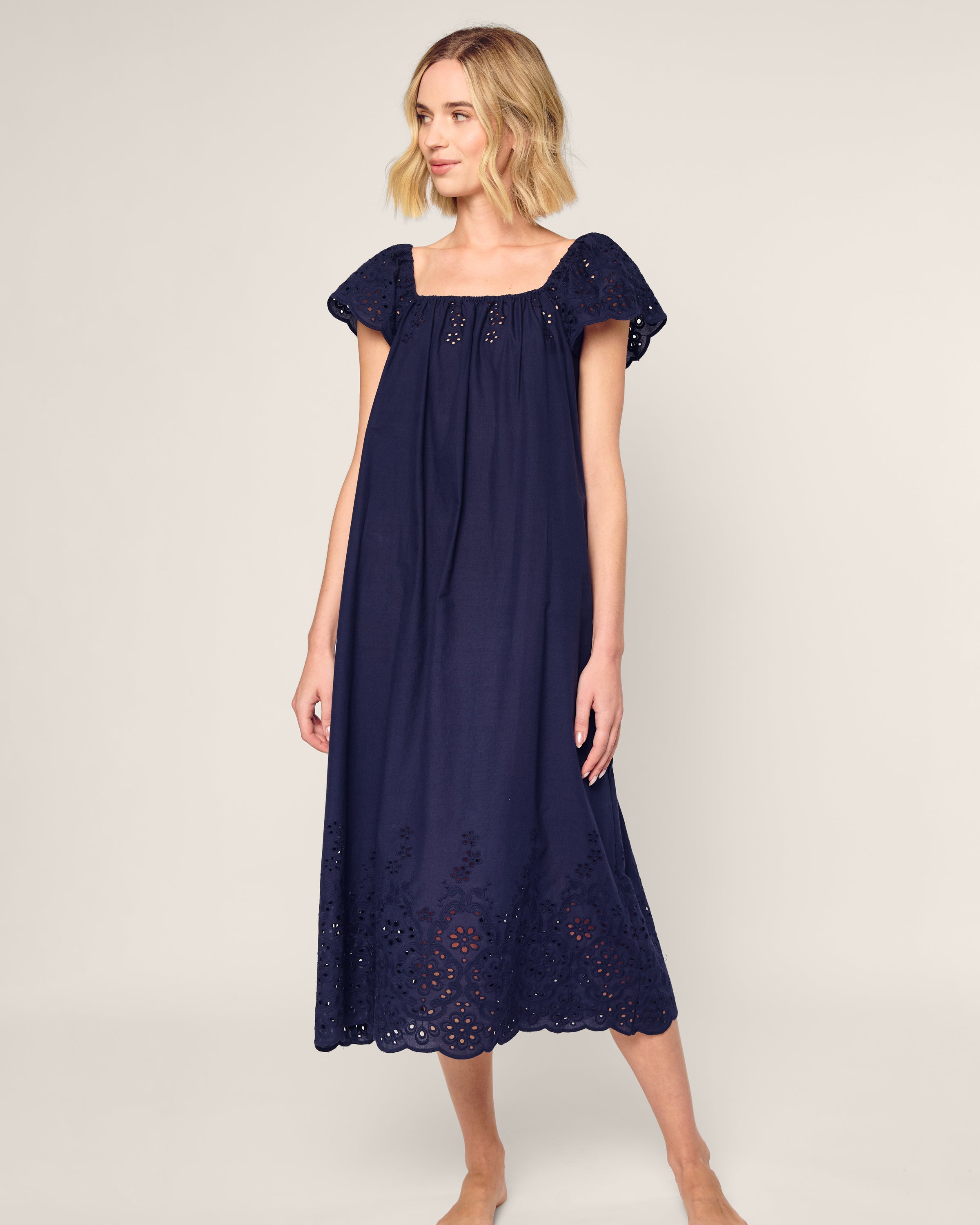 Barefoot, a person wears the Womens Twill Ava Nightgown in Navy by Petite Plume. This navy blue dress features short sleeves, eyelet details at the hem and sleeves, a square neckline, and a flowing silhouette. The background is plain and light-colored.
