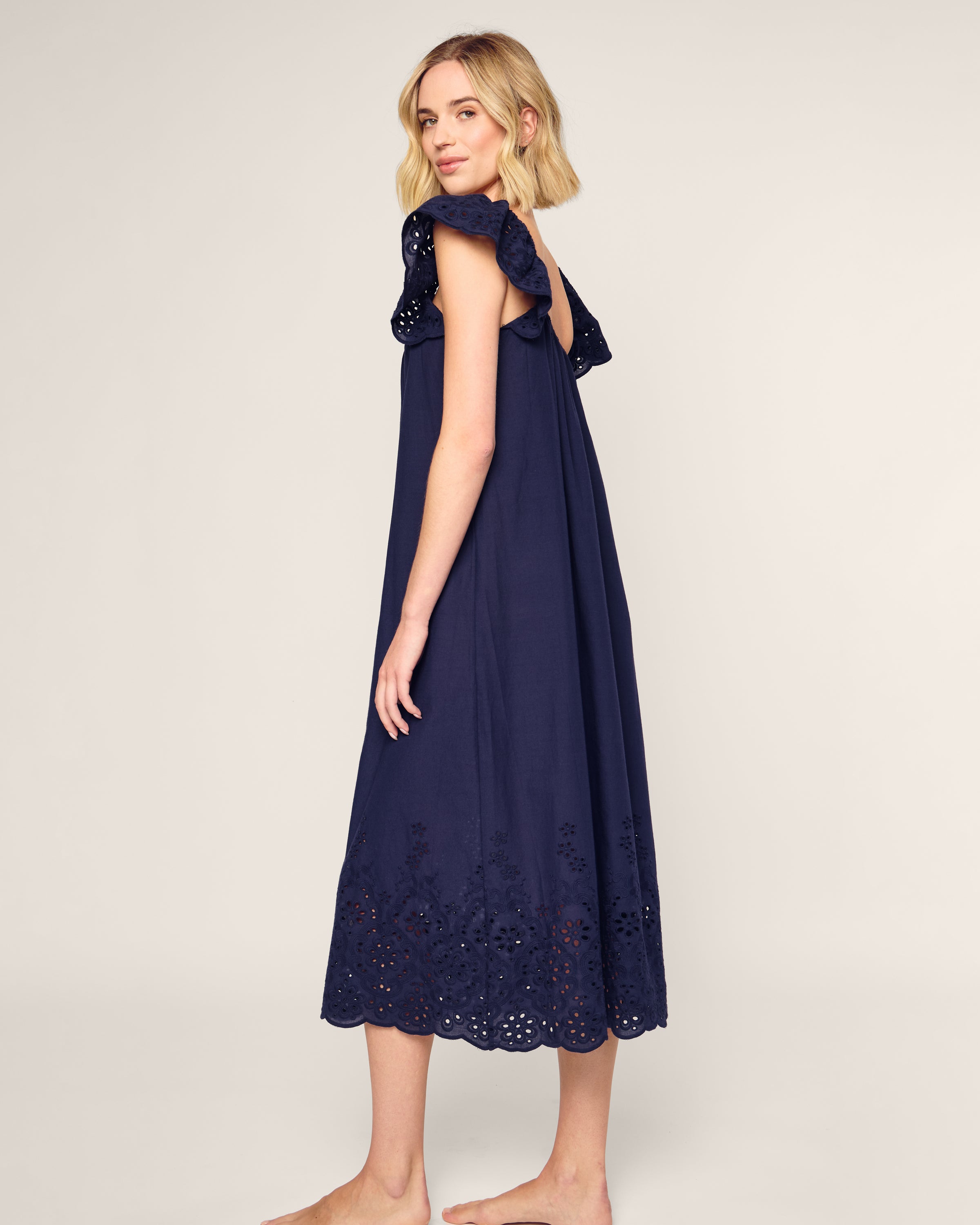 A barefoot woman with shoulder-length blonde hair stands sideways in a sleeveless navy blue dress with floral embroidery at the hem, gazing over her shoulder. She embodies the elegance of luxury sleepwear reminiscent of Petite Plumes Womens Twill Ava Nightgown in Navy against a plain backdrop.