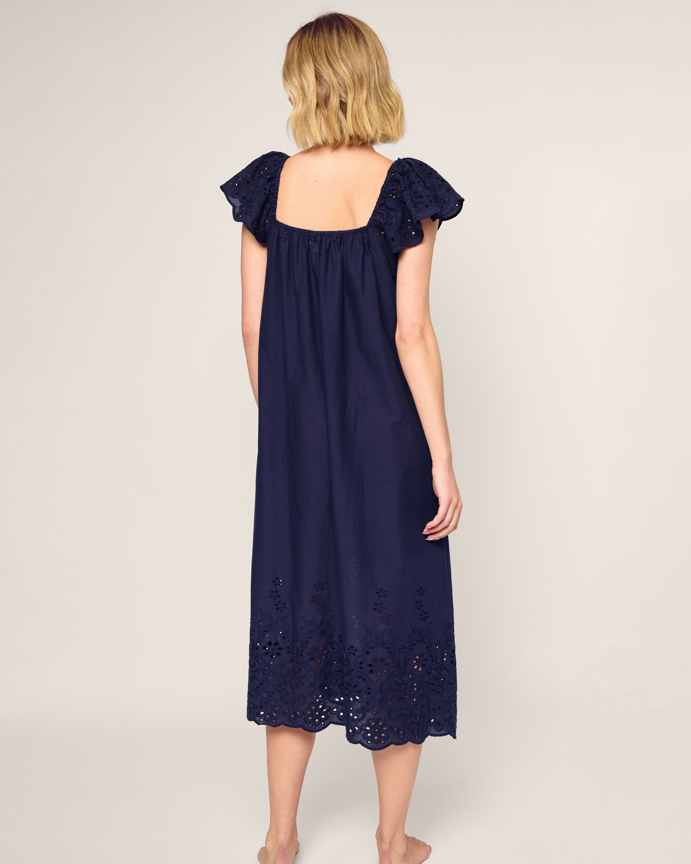 A person with shoulder-length blonde hair wears the Womens Twill Ava Nightgown in Navy by Petite Plume. This luxury sleepwear-inspired piece has flutter sleeves and eyelet hem detailing. Barefoot, they face away from the camera against a neutral background.