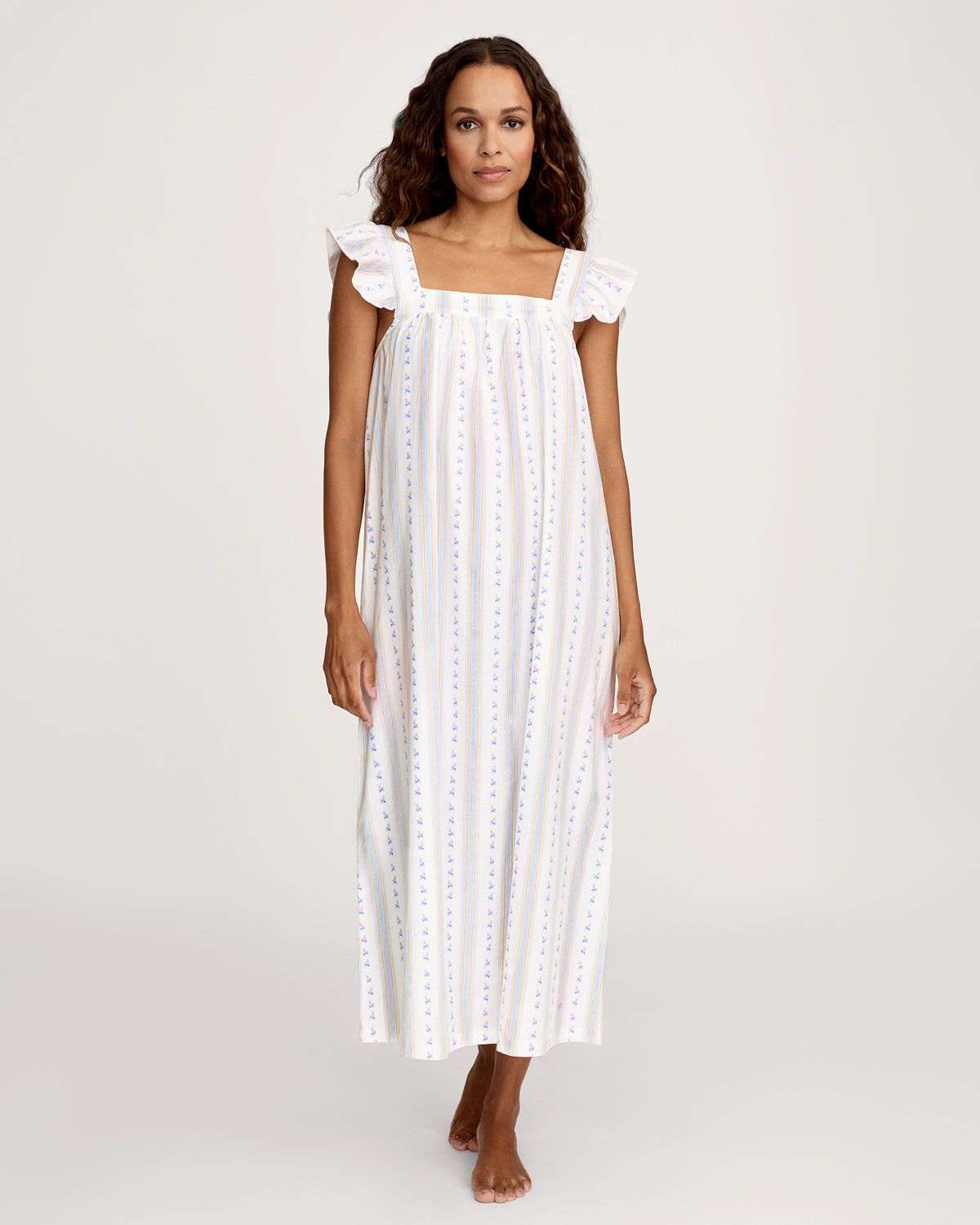 A woman stands barefoot against a plain light background wearing the Women's Twill Clara Nightgown in Cottage Stripe, featuring a long, loose fit with pastel vertical stripes and ruffled shoulder straps.