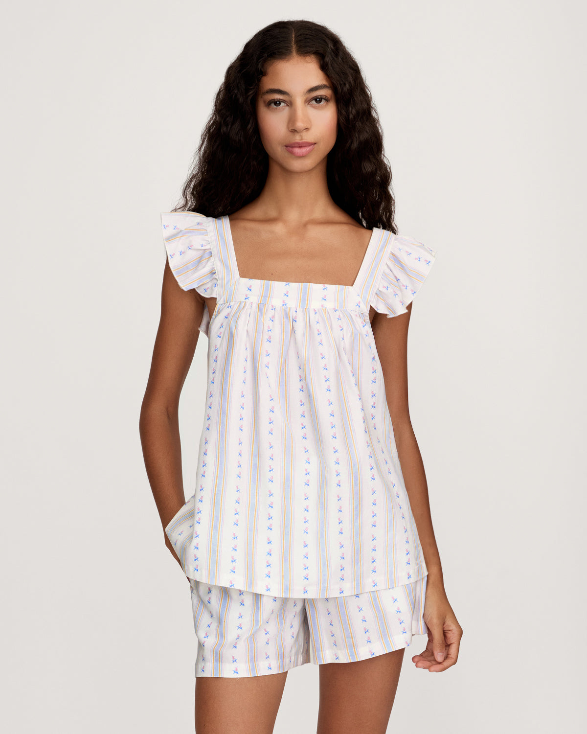 A woman with long curly hair models the Women's Twill Clara Short Set in Cottage Stripe—a loose, sleeveless pajama set with ruffled shoulders and shorts, featuring white fabric with pastel stripes and floral patterns—against a plain background.
