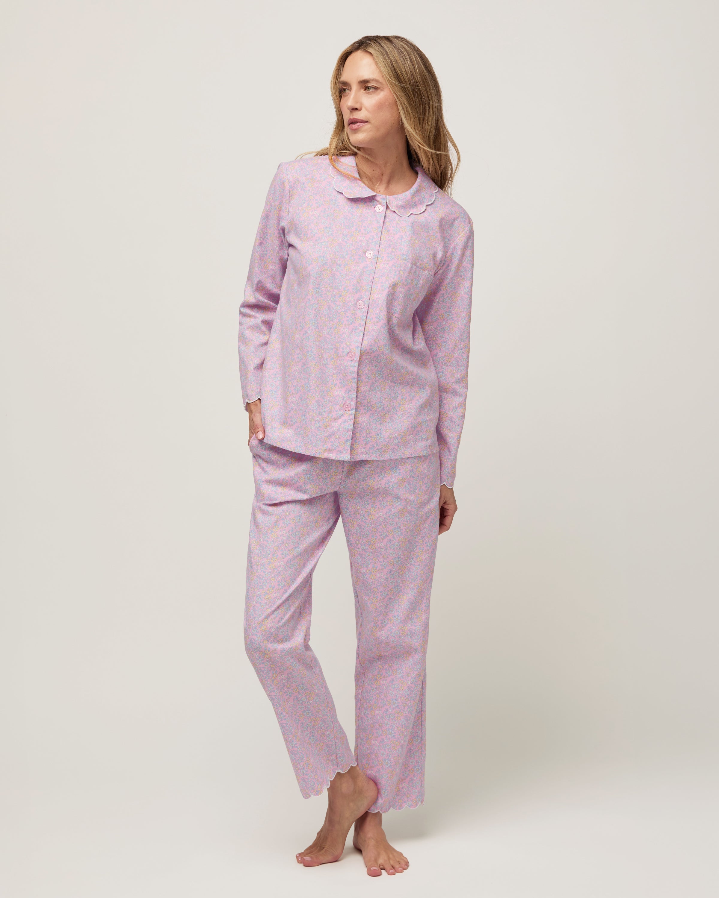 A woman stands barefoot against a plain background, wearing the Women's Twill Colette Pajama Set in Fleur Blush—a matching long-sleeve, button-up top and pants with a light purple and pink floral pattern.