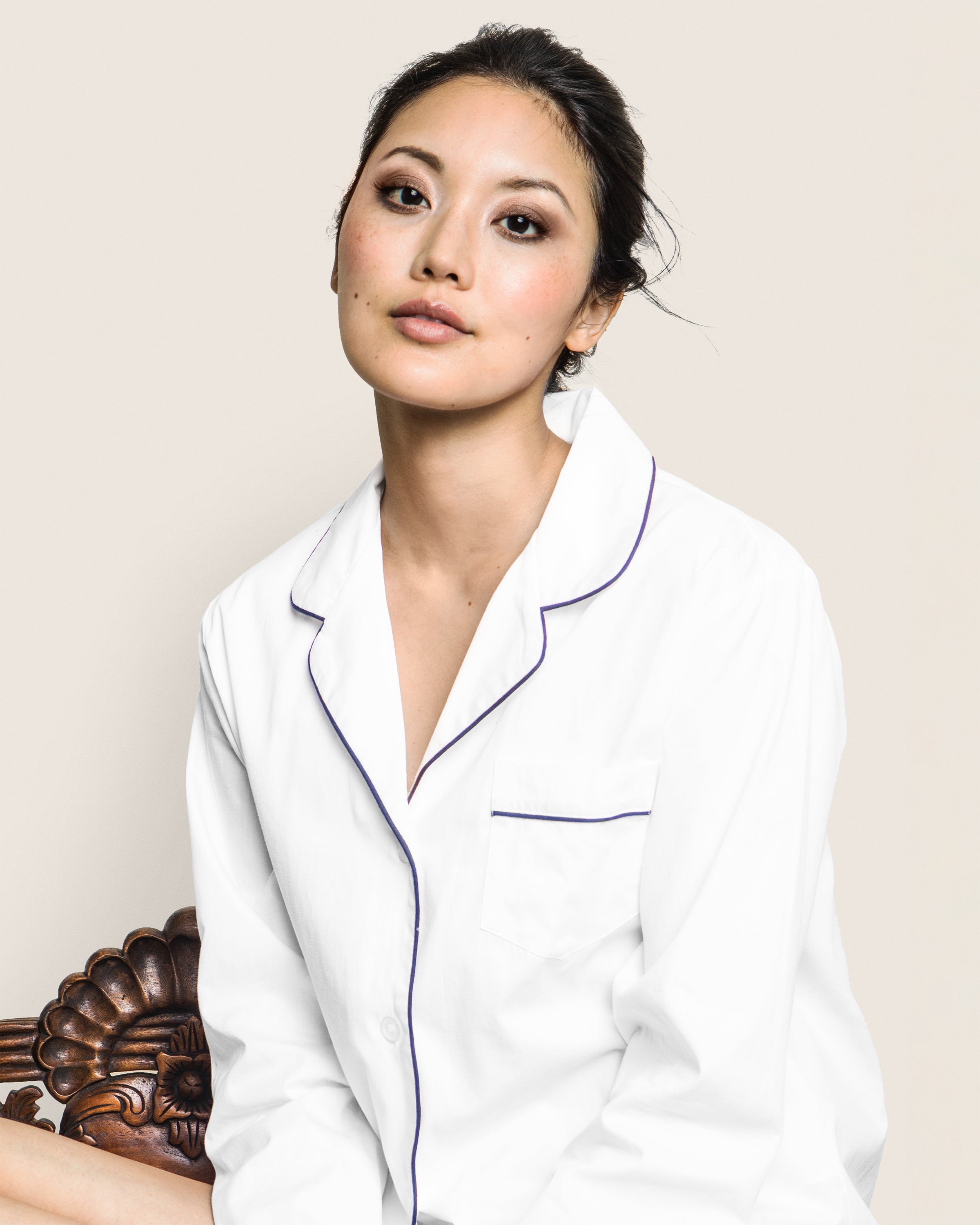 A calm person wearing Petite Plumes Womens Long Sleeve Short Set in White Twill with Navy Piping is seated against an ornate wooden chair. The background is neutral, and their dark hair is tied back.