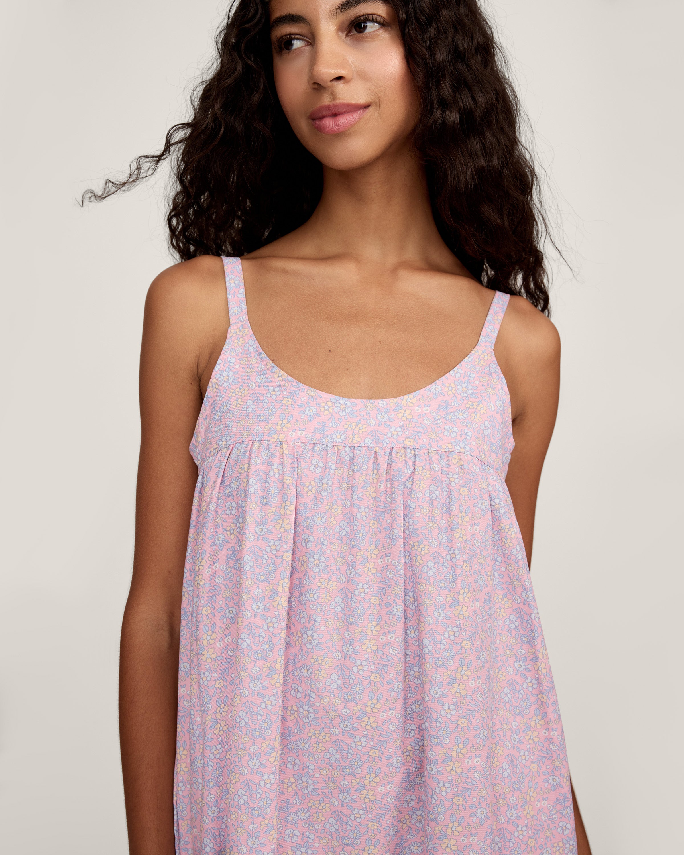 A woman with long curly hair models the Women's Twill Chloé Nightgown in Fleur Blush, a sleeveless, loose-fitting light purple floral dress. She is barefoot and stands relaxed against a plain, light background.