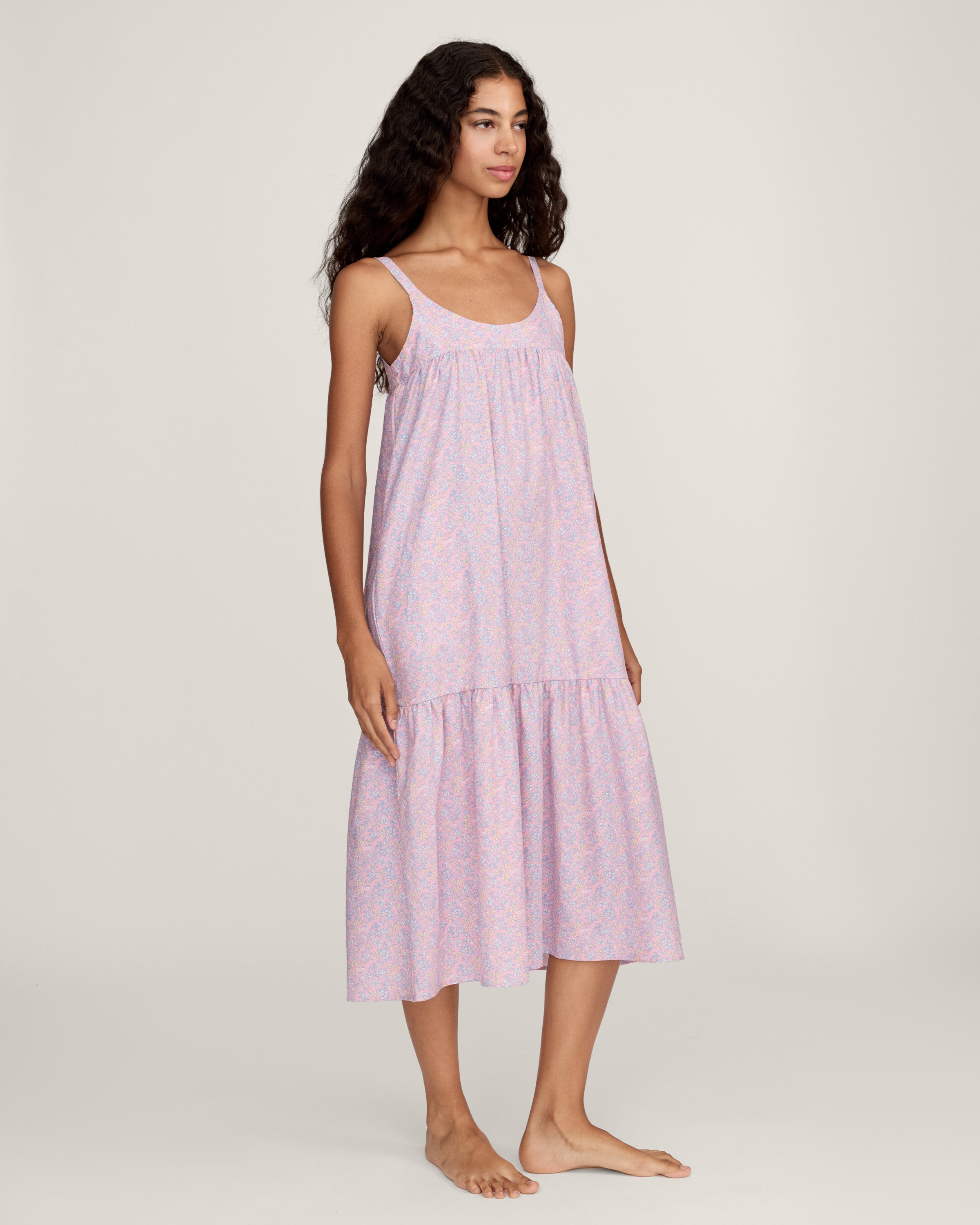 A woman with long curly hair models the Women's Twill Chloé Nightgown in Fleur Blush, a sleeveless, loose-fitting light purple floral dress. She is barefoot and stands relaxed against a plain, light background.