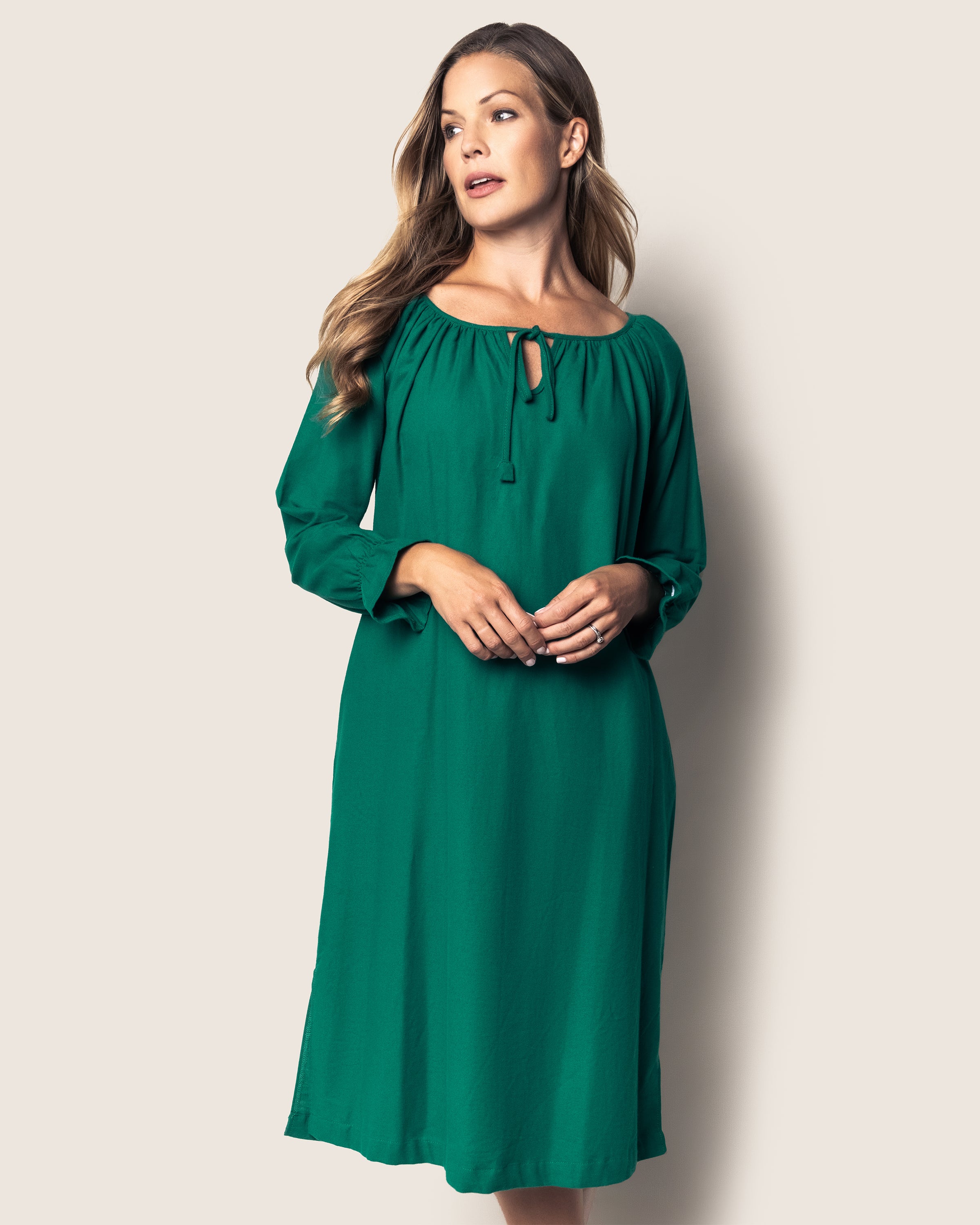 A woman with long hair wears the Womens Green Flannel Delphine Nightgown by Petite Plume, resembling an elegant, green knee-length dress with long sleeves and a keyhole neckline. She gazes sideways with her hands clasped against a plain light background.