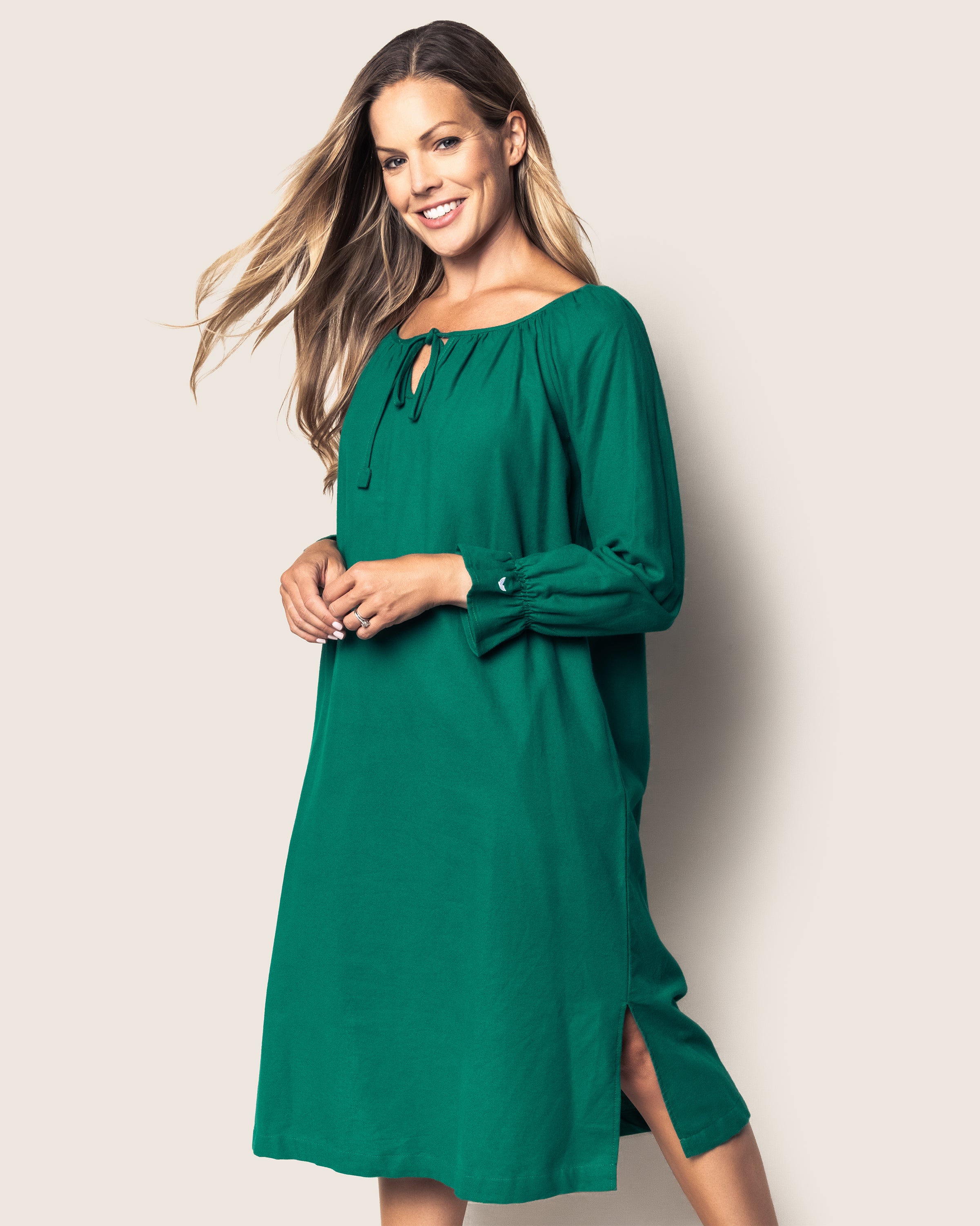 A smiling woman wears the Womens Green Flannel Delphine Nightgown by Petite Plume, featuring a tied neckline and side slits. Her long hair cascades over her shoulders as she stands against a neutral background.
