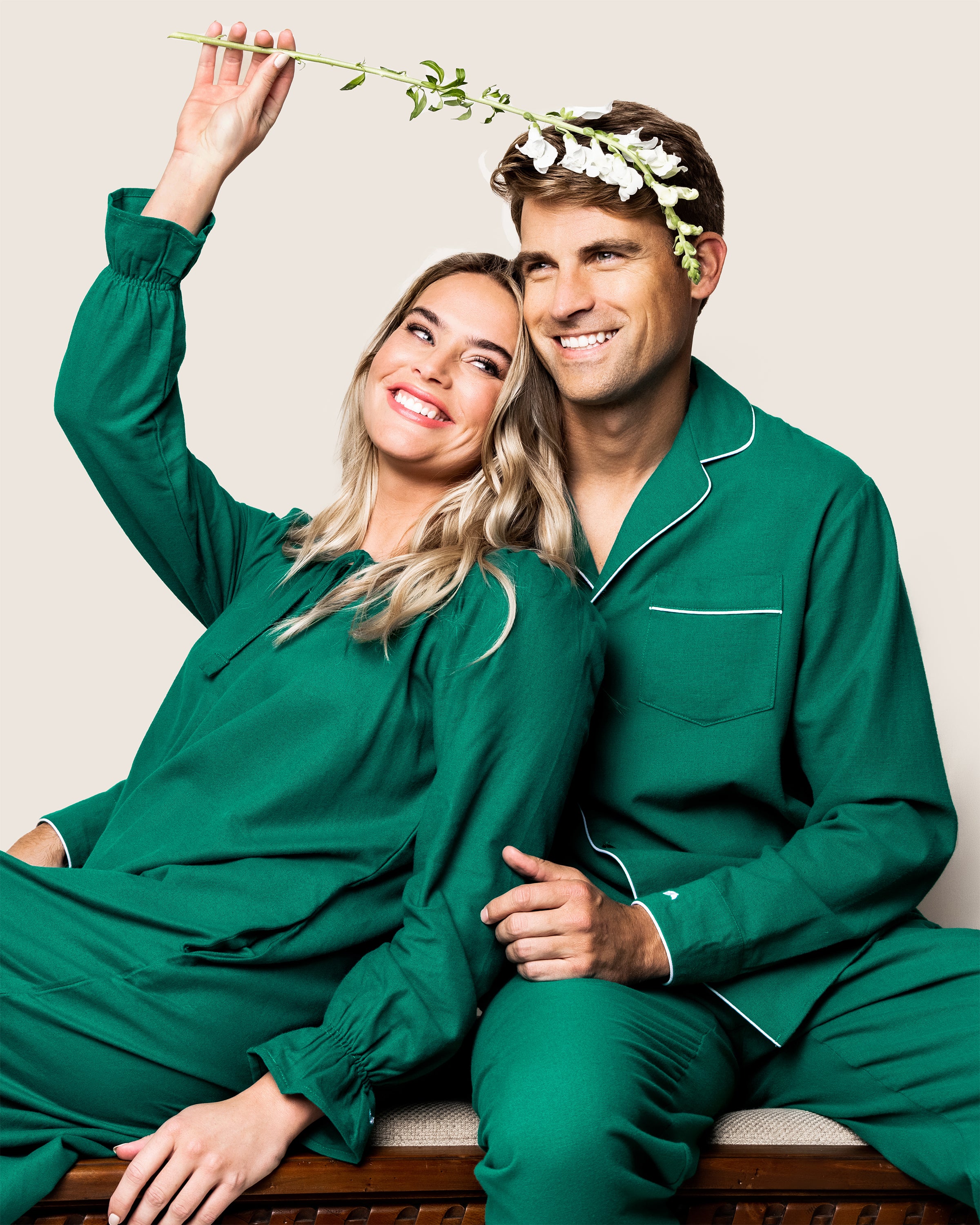 A smiling woman, wearing a Womens Green Flannel Delphine Nightgown by Petite Plume, and a man sit together in matching green brushed cotton pajamas. She playfully holds a sprig of white flowers above them. The background is a simple off-white.