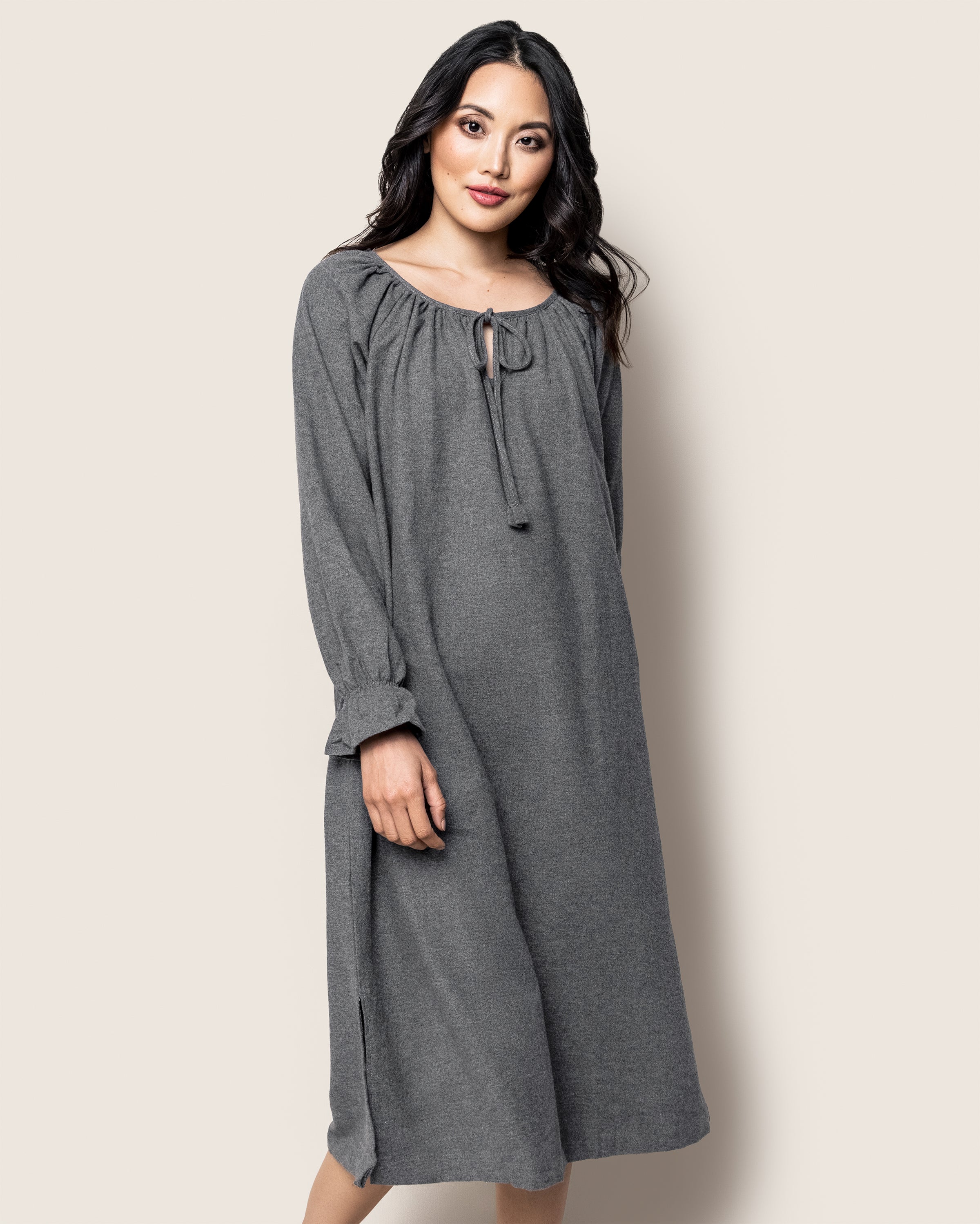 Against a plain background, a woman poses in the Womens Flannel Delphine Nightgown by Petite Plume. Its gray with long sleeves, made of 100% cotton soft flannel, and features a drawstring neckline and loose fit. She stands with one hand on her hip, smiling softly.