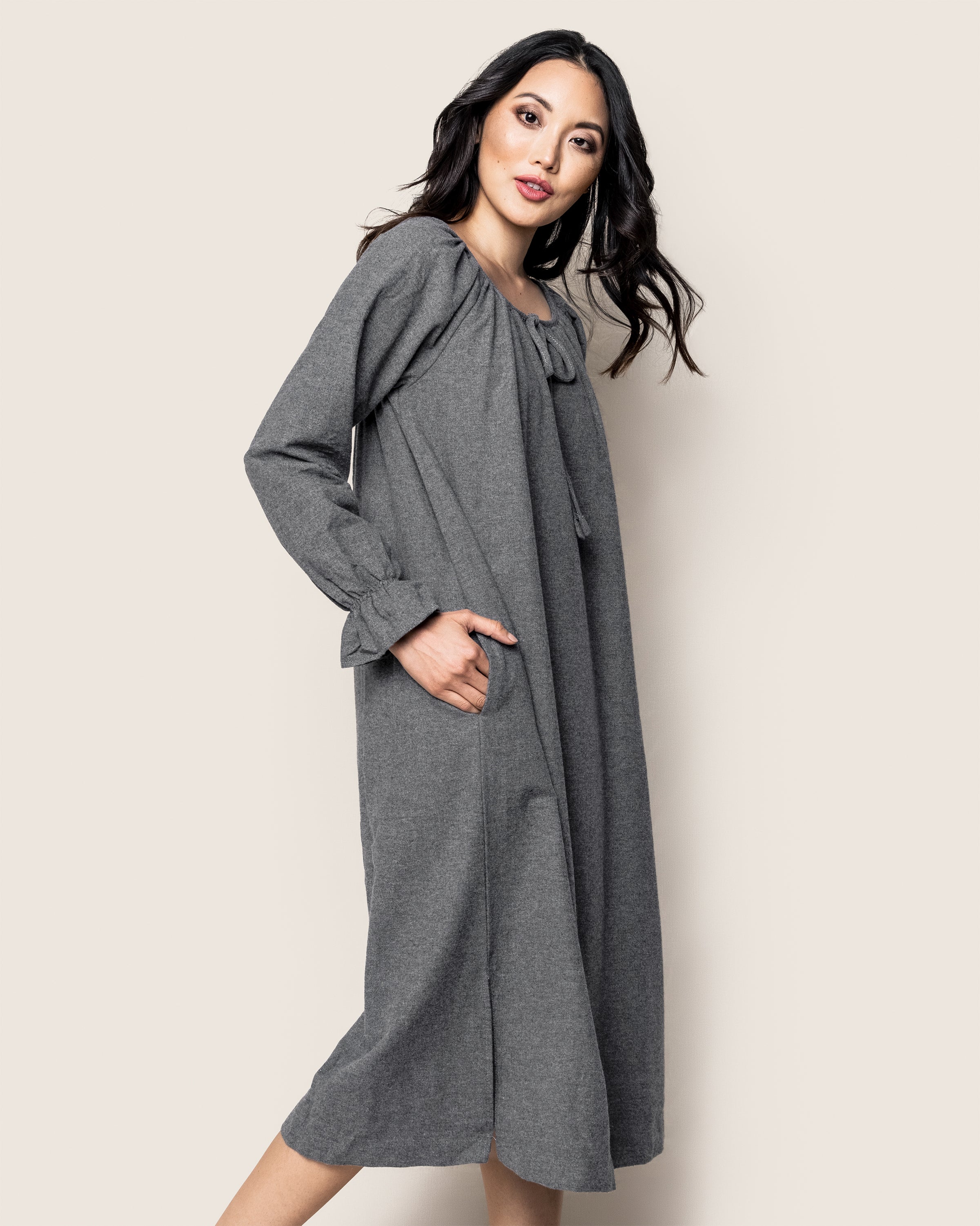 The woman with long dark hair wears a loose-fitting, 100% cotton Womens Flannel Delphine Nightgown in Grey by Petite Plume, featuring pockets. She stands confidently against a plain light background, embodying relaxed elegance while looking over her shoulder.