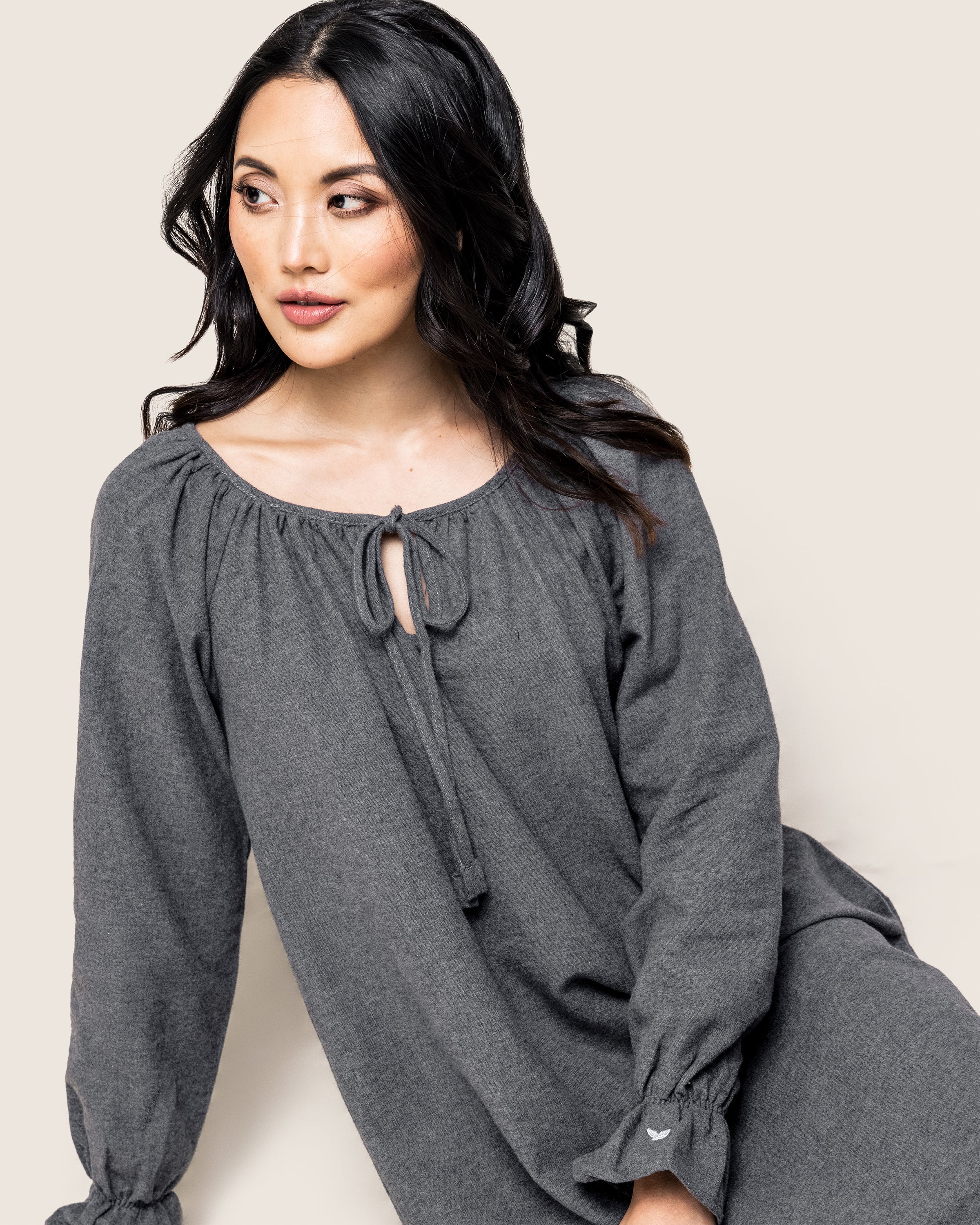A woman with long dark hair models Petite Plumes Womens Flannel Delphine Nightgown in Grey. Featuring long sleeves and a tie at the neckline, she exudes relaxed elegance against a light beige background.