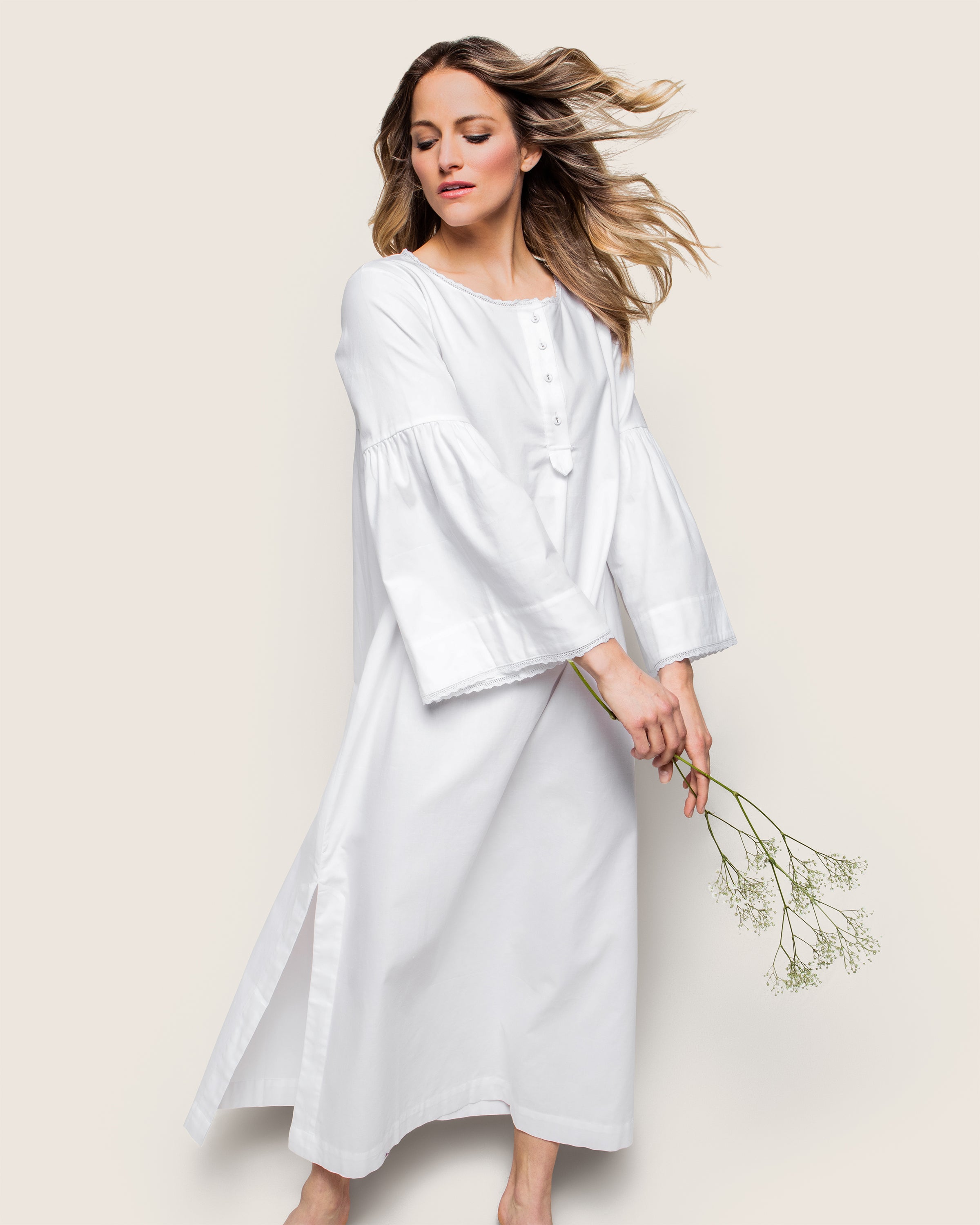 A woman in a long, Petite Plume Womens Flannel Seraphine Nightgown in White stands against a beige background, her hair flowing and holding delicate white flowers. She radiates serenity and grace with the finest quality cotton fabric.