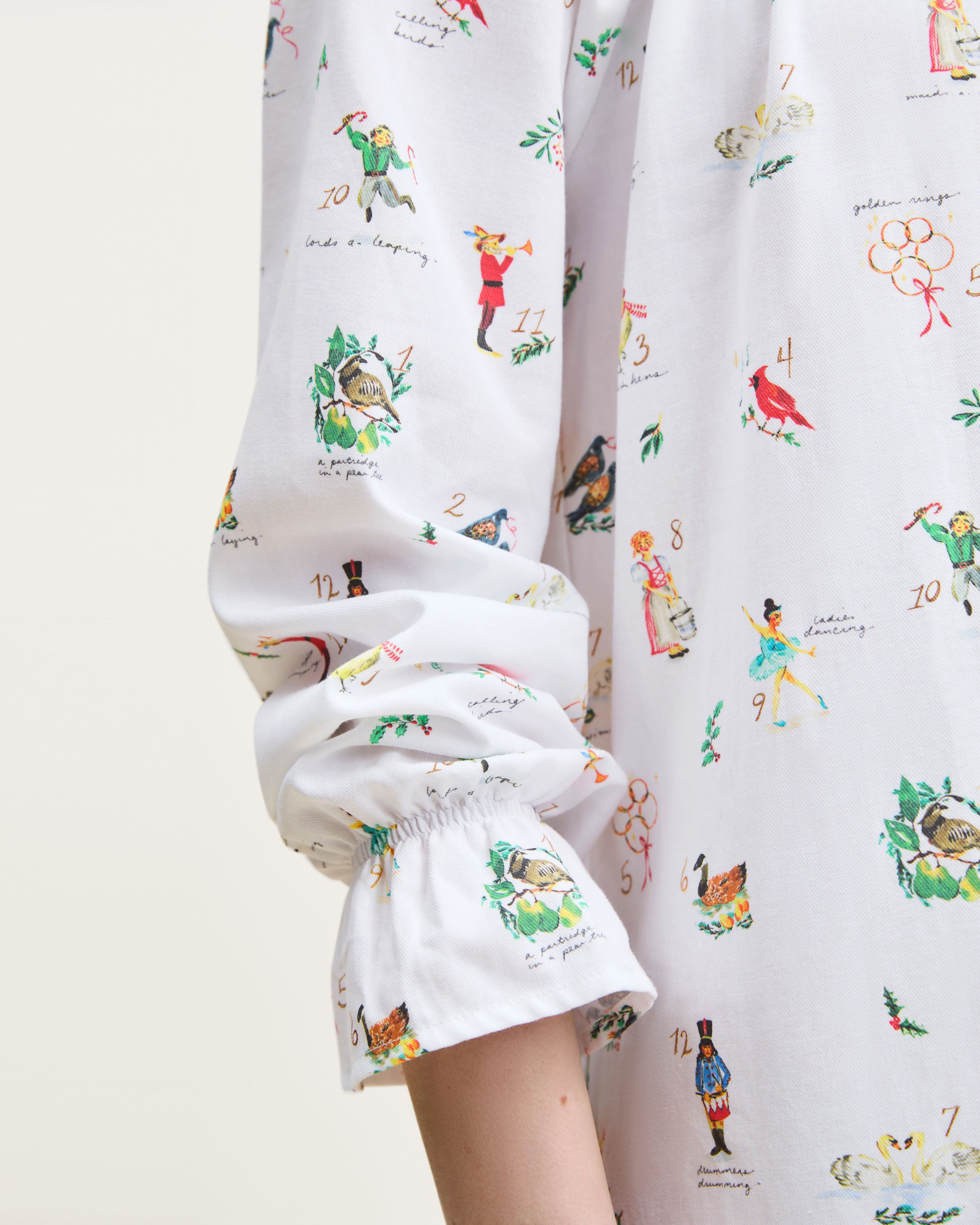 The Women's Twill Delphine Nightgown in 12 Days of Christmas is a white, long-sleeve nightgown with a gathered neckline, small bow tie, and a festive print inspired by the 12 Days of Christmas.