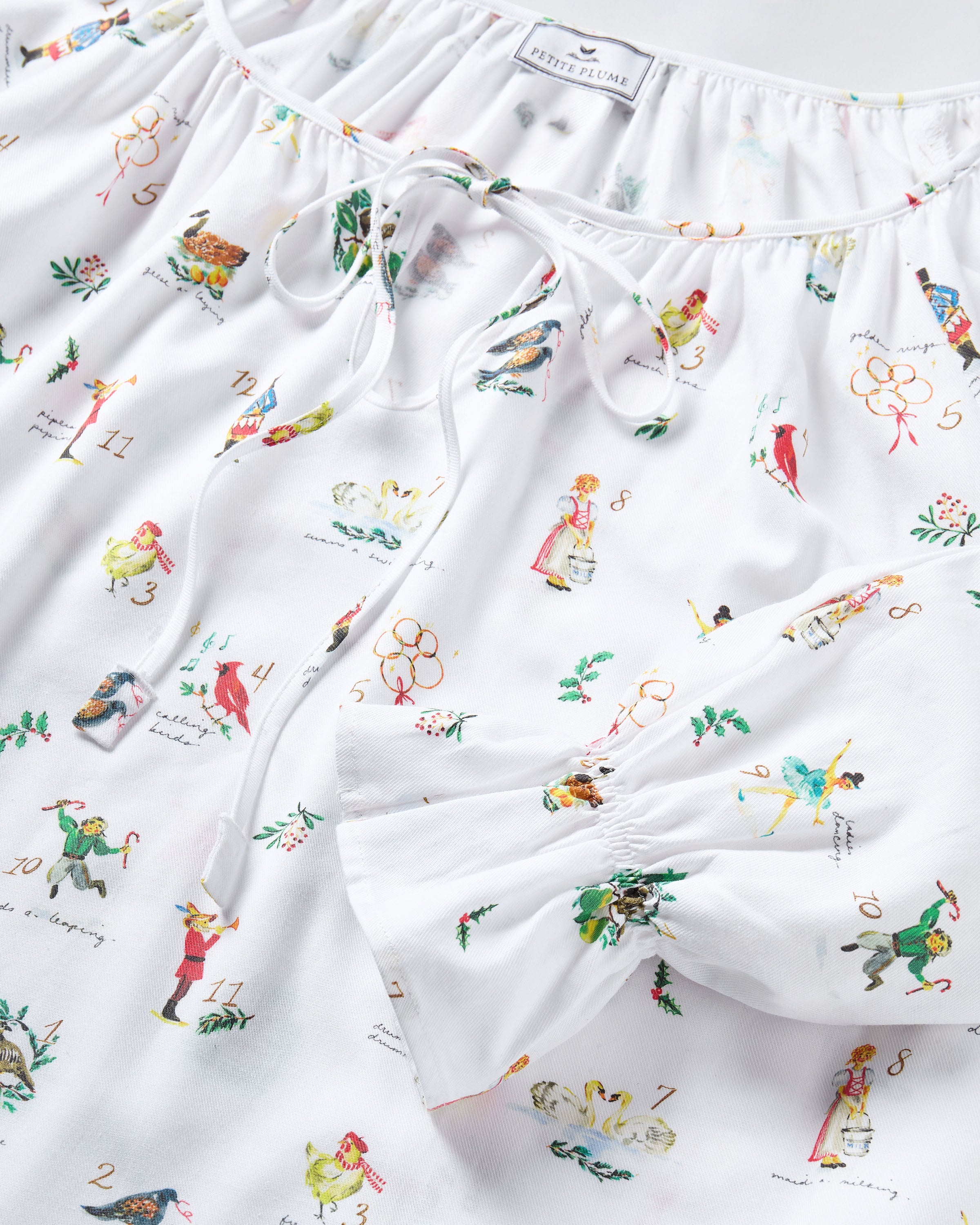 The Women's Twill Delphine Nightgown in 12 Days of Christmas is a white, long-sleeve nightgown with a gathered neckline, small bow tie, and a festive print inspired by the 12 Days of Christmas.
