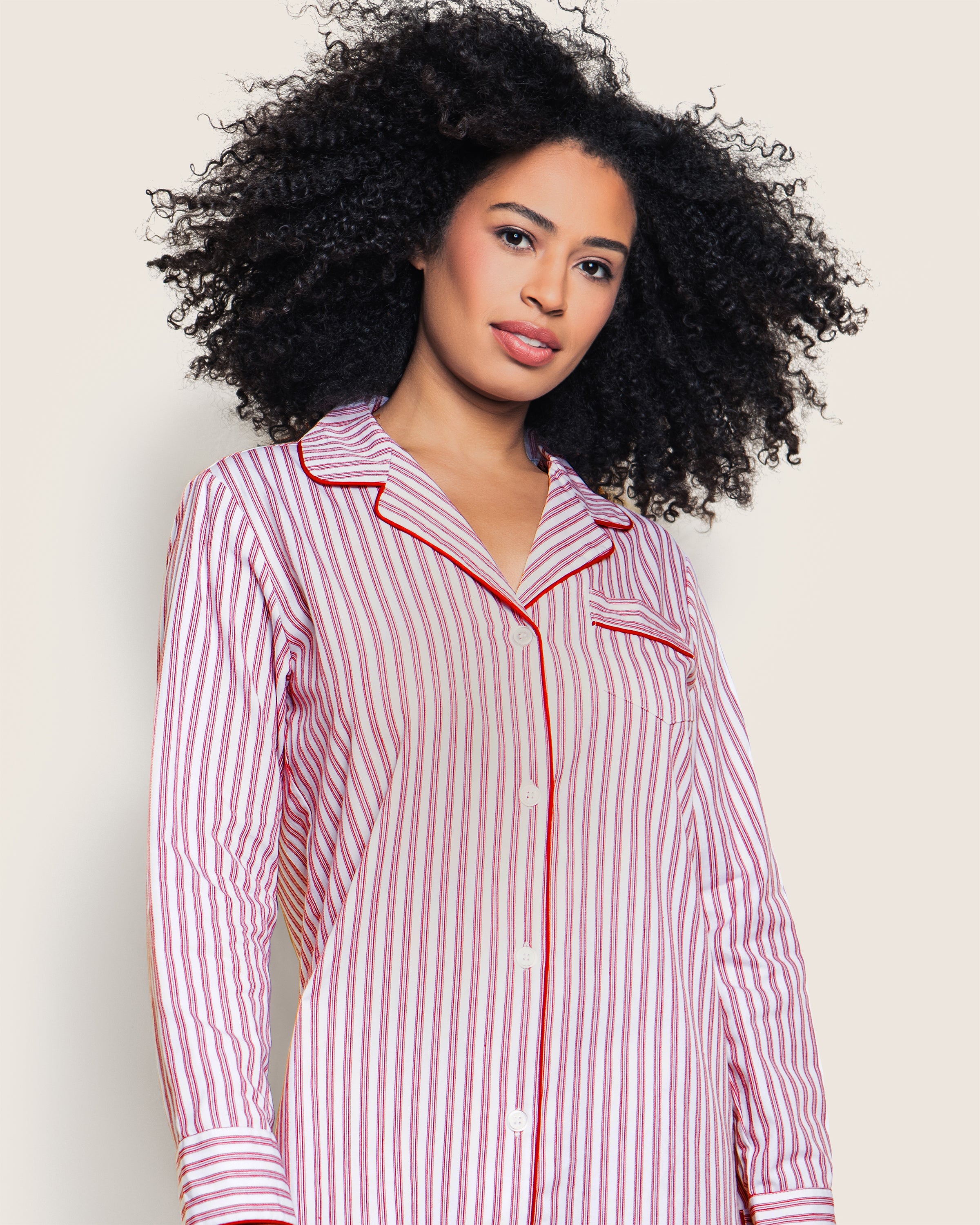 A curly-haired person smiles, wearing Petite Plumes Womens Twill Nightshirt in Antique Red Ticking, featuring a collar and a breast pocket. The pink and white striped button-up shirt is made of the finest quality cotton, while the plain background enhances its look.