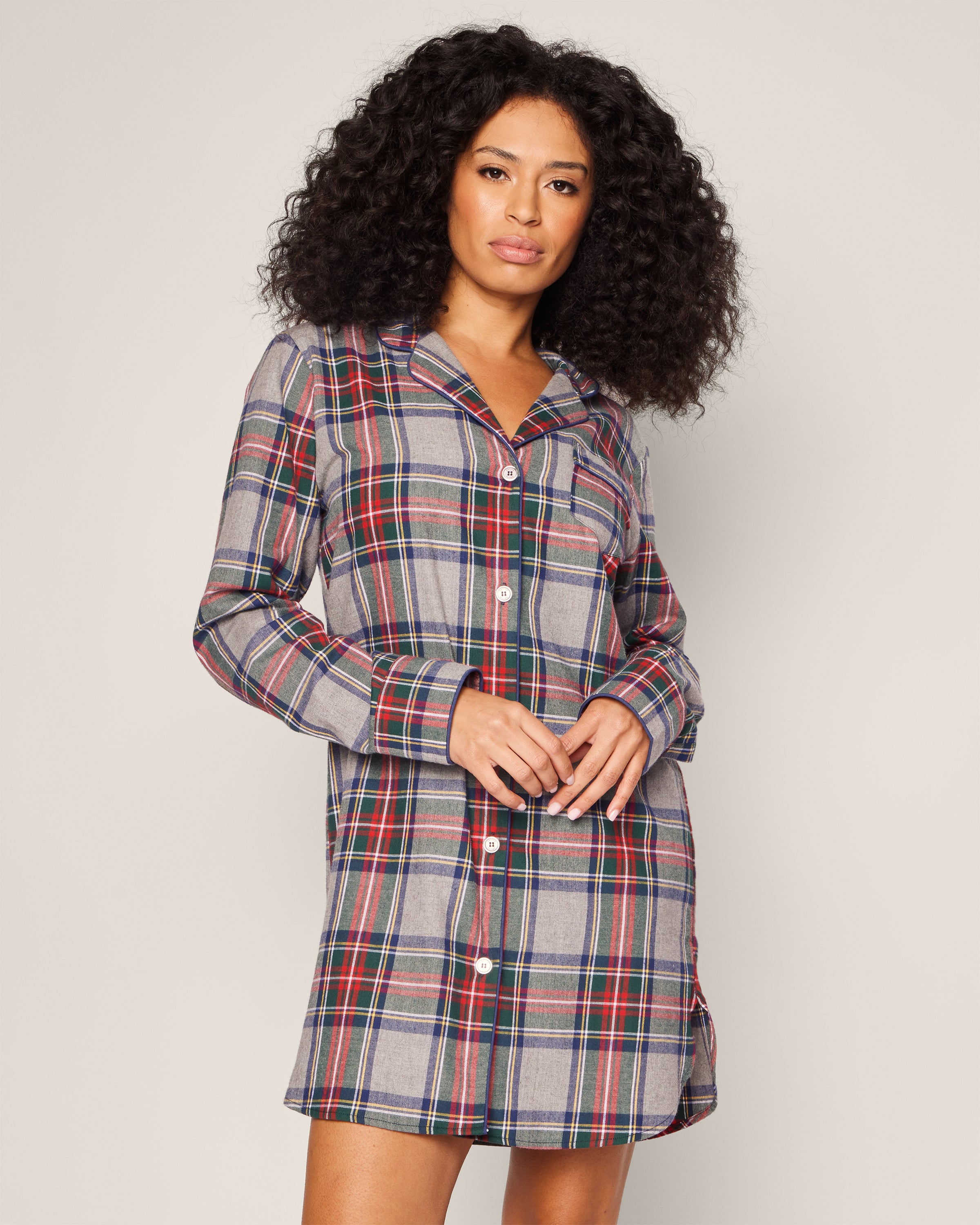 A person with curly hair wears Petite Plumes Womens Brushed Cotton Nightshirt in Westminster Tartan featuring rich red, blue, and green tones, standing against a plain background while looking at the camera.