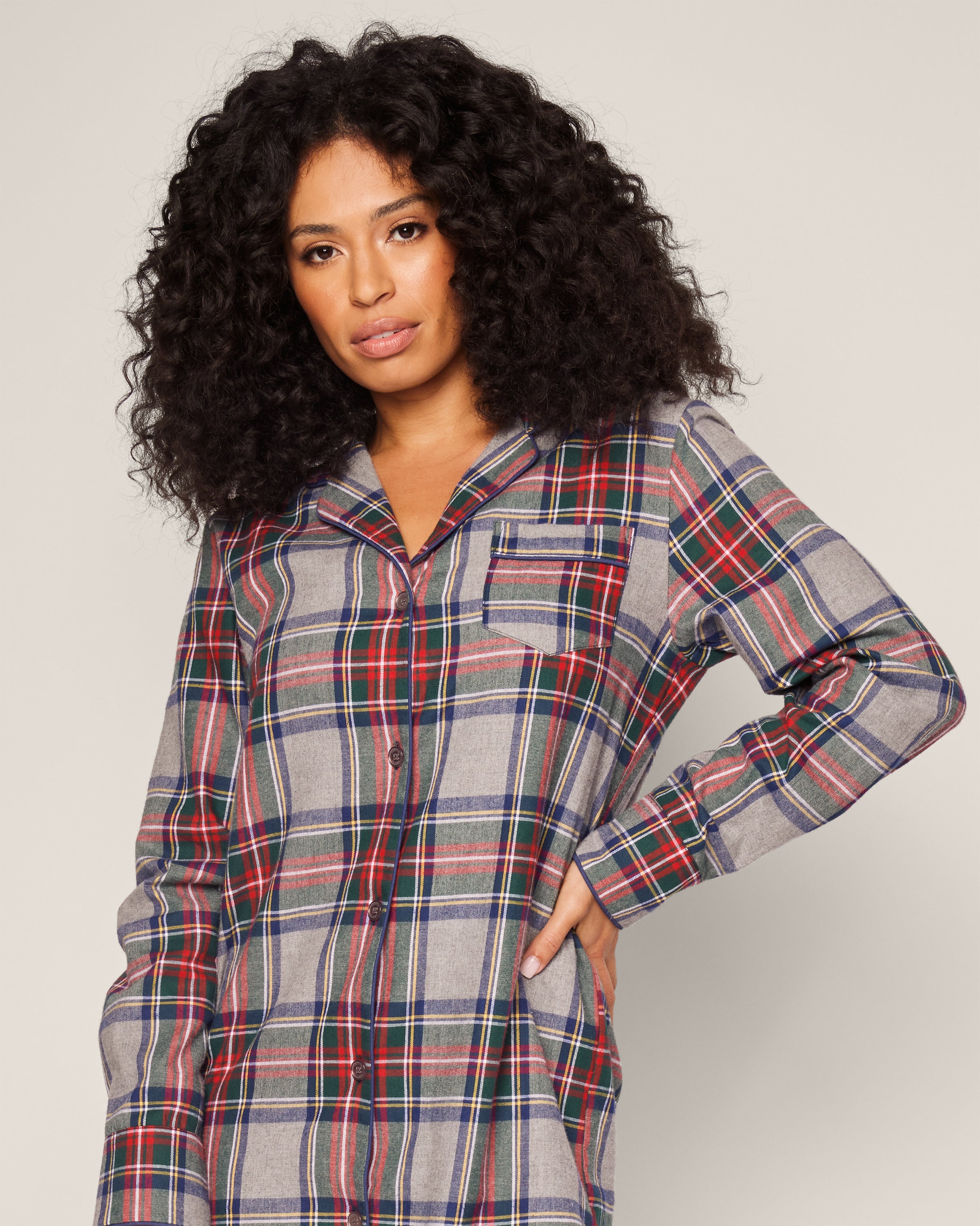 A person with curly hair sports the Womens Brushed Cotton Nightshirt by Petite Plume, showcasing Westminster Tartans rich patterns in red, blue, and gray. They stand relaxed against a plain backdrop, one hand casually in their pocket.
