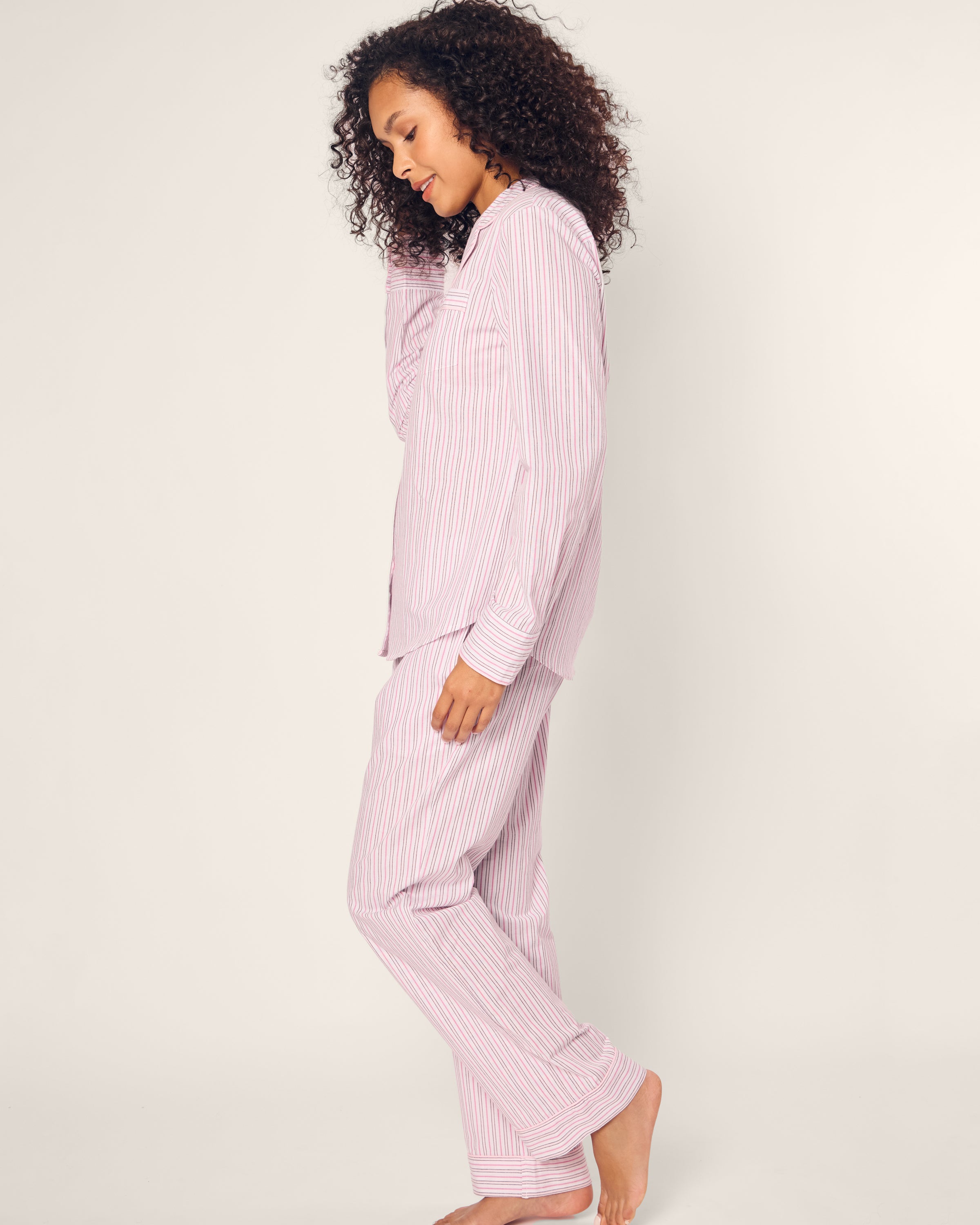 A person with curly hair stands sideways in Petite Plumes Womens Twill Pajama Set in Blush Boulevard, against an off-white background. Made from 100% cotton, they are barefoot and appear relaxed.