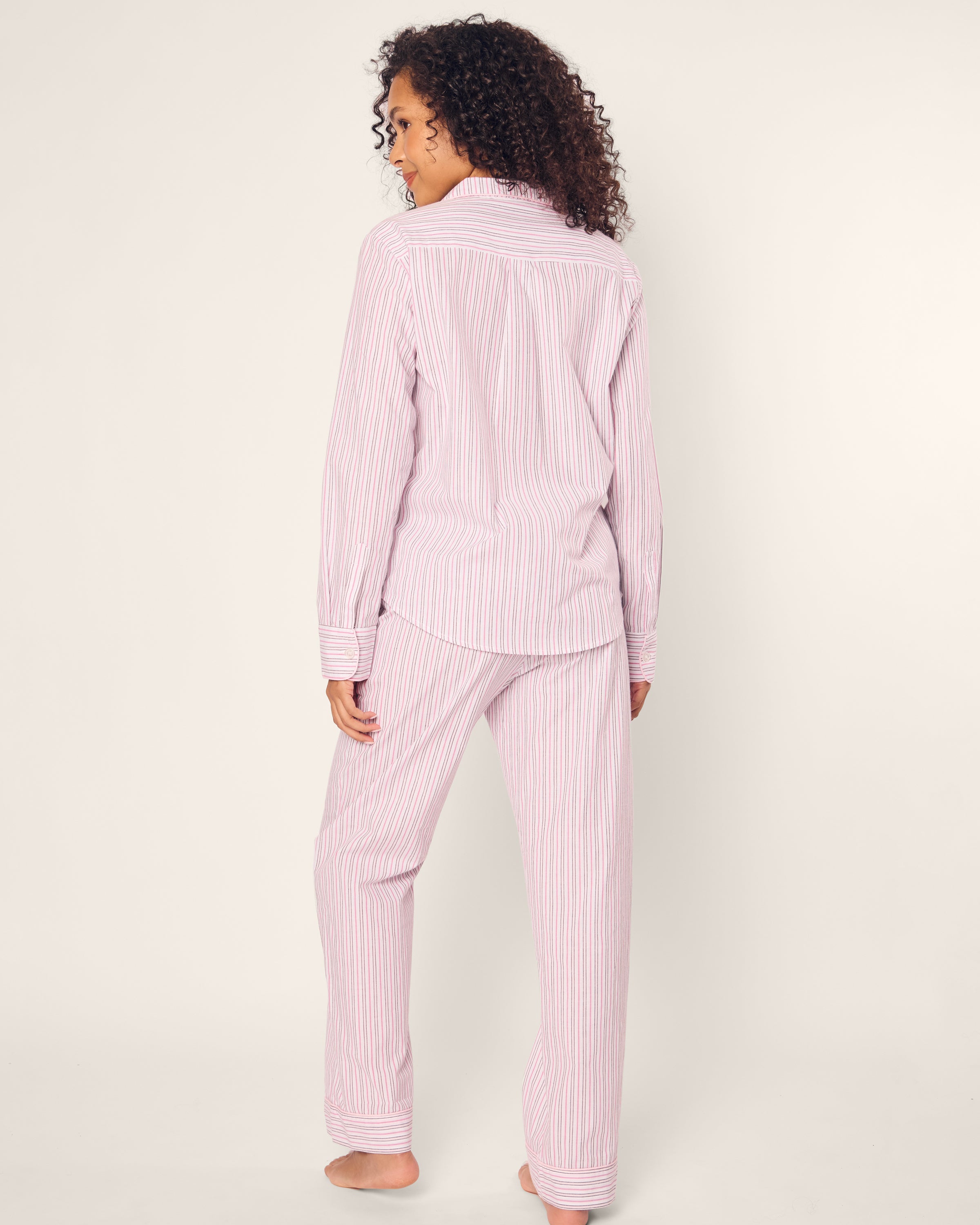 A person with curly hair stands barefoot, facing away, wearing Petite Plumes Womens Twill Pajama Set from Blush Boulevard. This classic set features high-quality cotton pink-and-white striped long-sleeve shirt and pants against a plain light background.