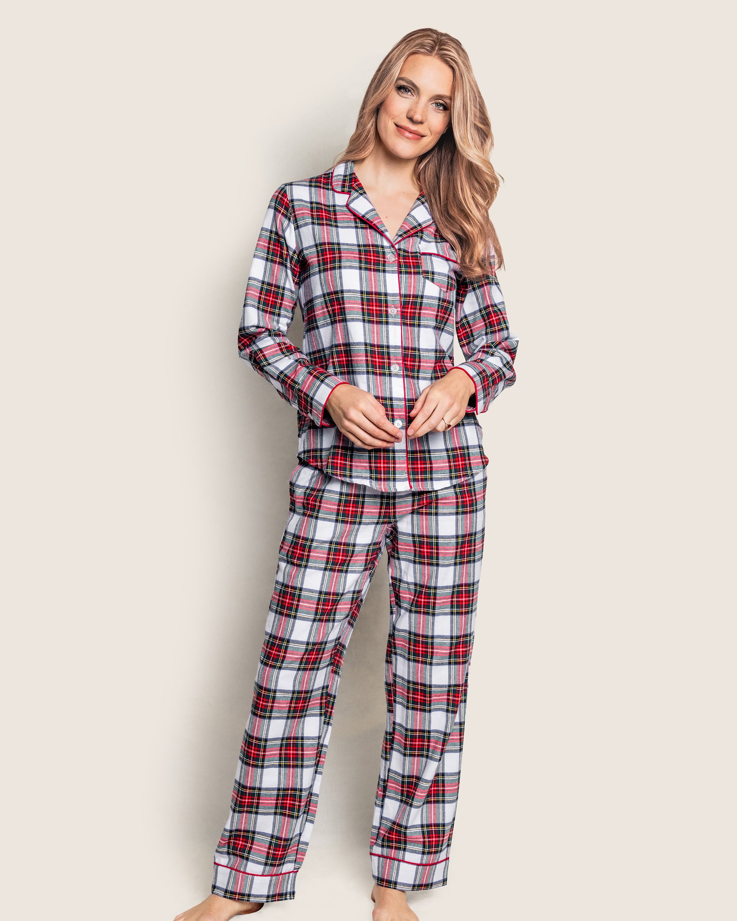 A person wearing Petite Plumes Womens Brushed Cotton Pajama Set in Balmoral Tartan stands barefoot, smiling with hands lightly clasped, against a plain background.