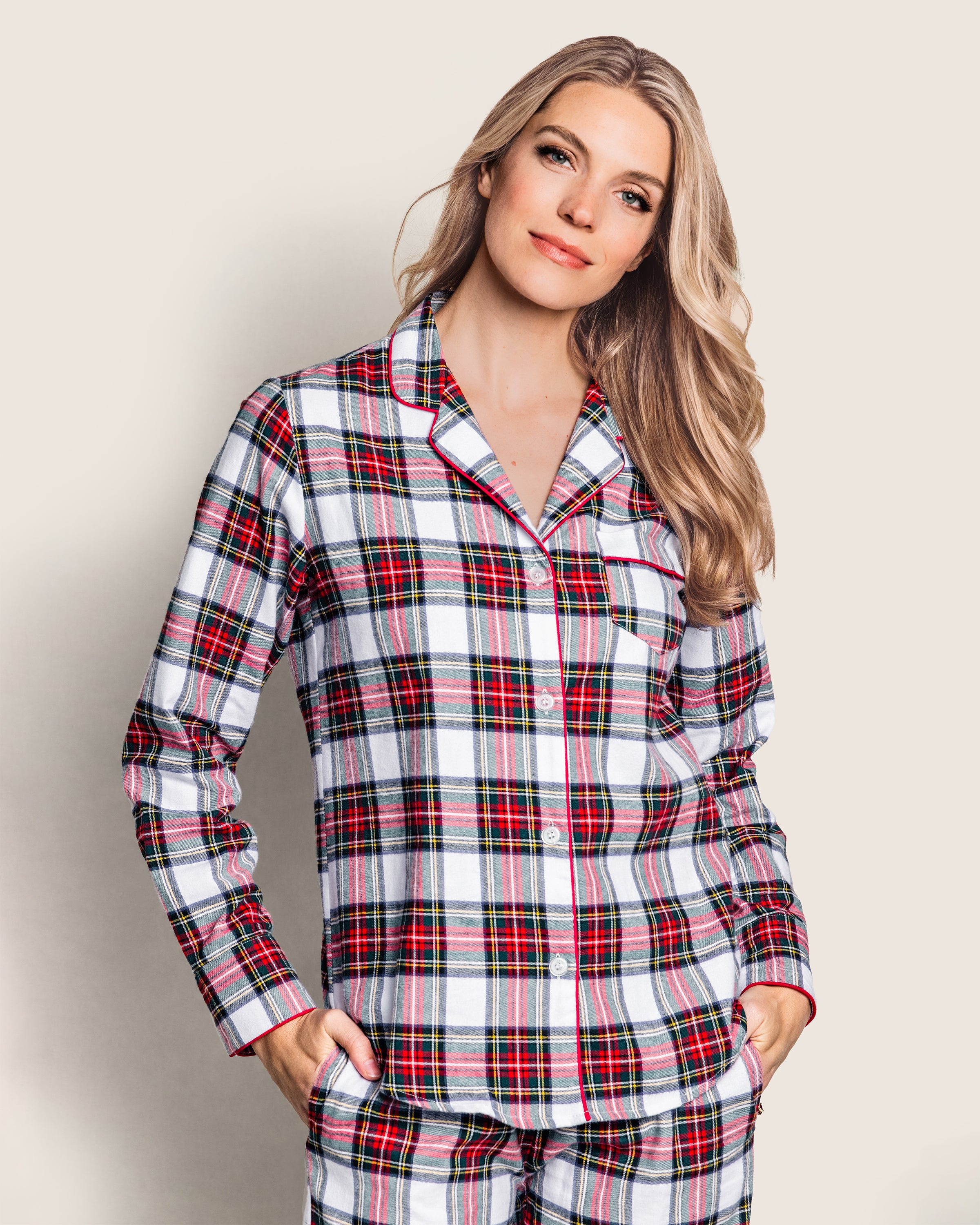 A long-haired blonde is wearing the Petite Plume Womens Brushed Cotton Pajama Set in Balmoral Tartan, featuring red, black, and green on white. Standing against a light backdrop with hands in pockets, they smile slightly.