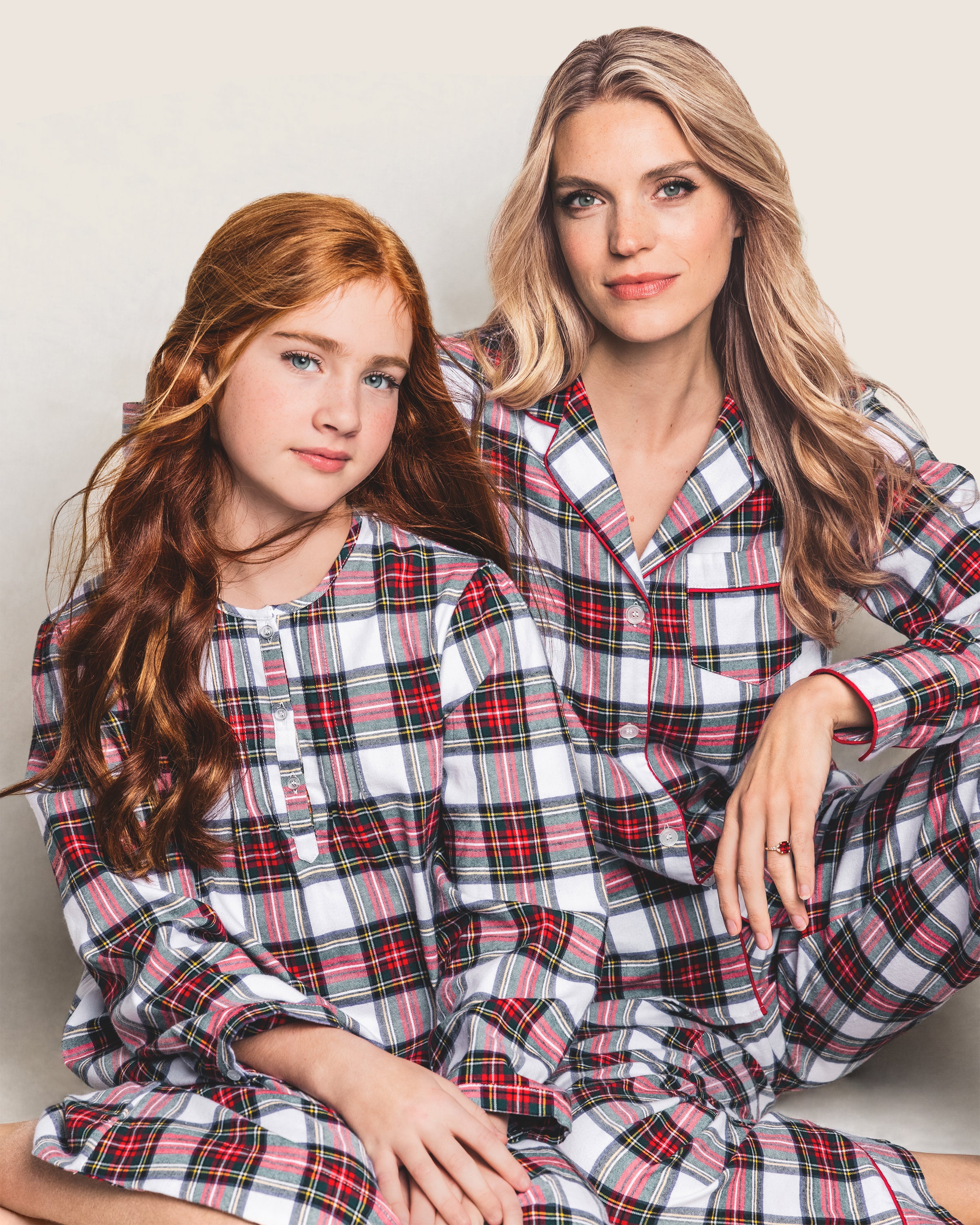 Two people in matching Womens Brushed Cotton Pajama Sets in Balmoral Tartan by Petite Plume sit together. One has long red hair, the other long blonde. They appear relaxed, smiling gently against a plain background.