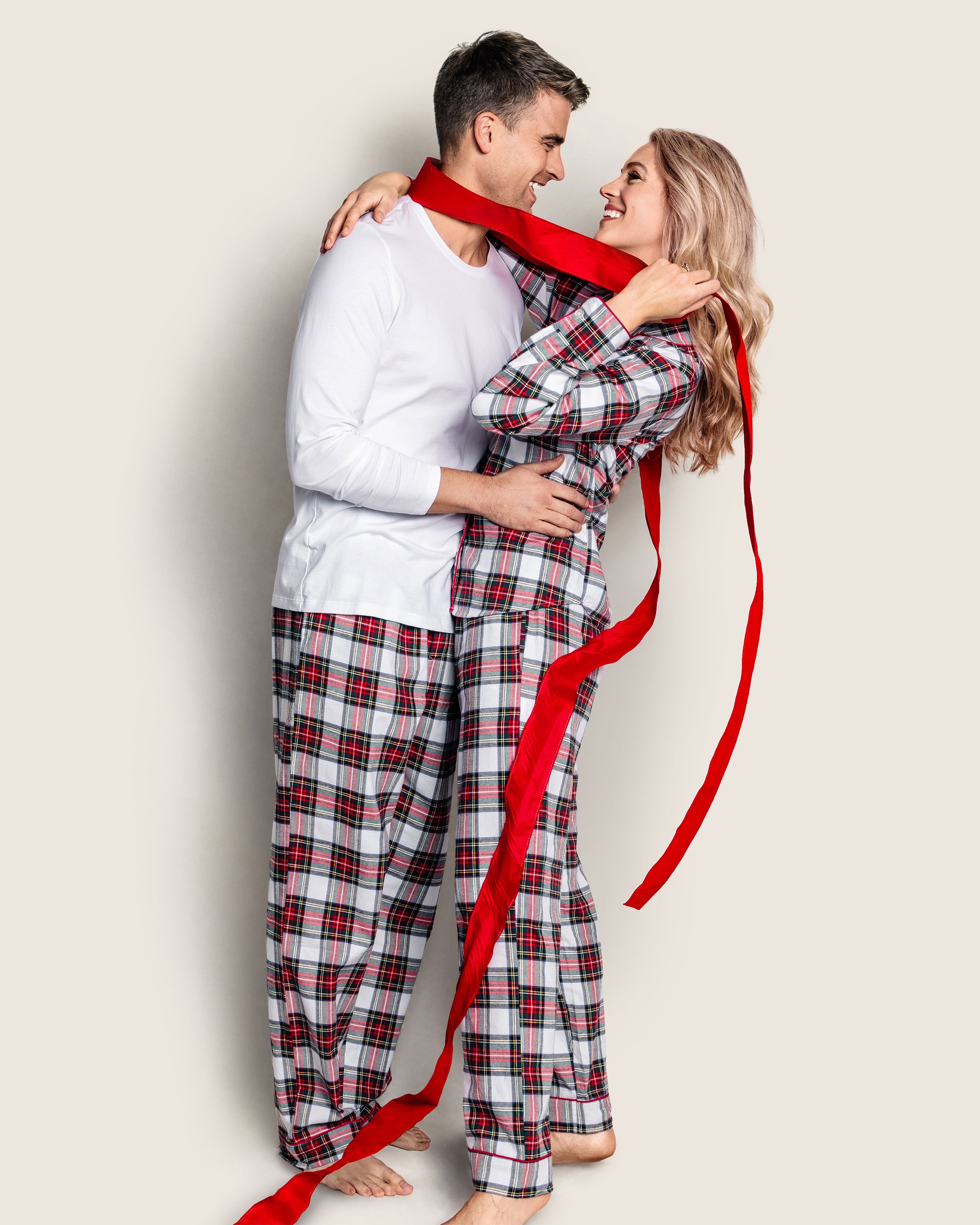 A couple in matching Petite Plume pajamas, featuring the womans Brushed Cotton Pajama Set in Balmoral Tartan, stands closely smiling. The man wears a white long-sleeve shirt while she playfully wraps herself in a red ribbon, adding charm against the plain light-colored background.