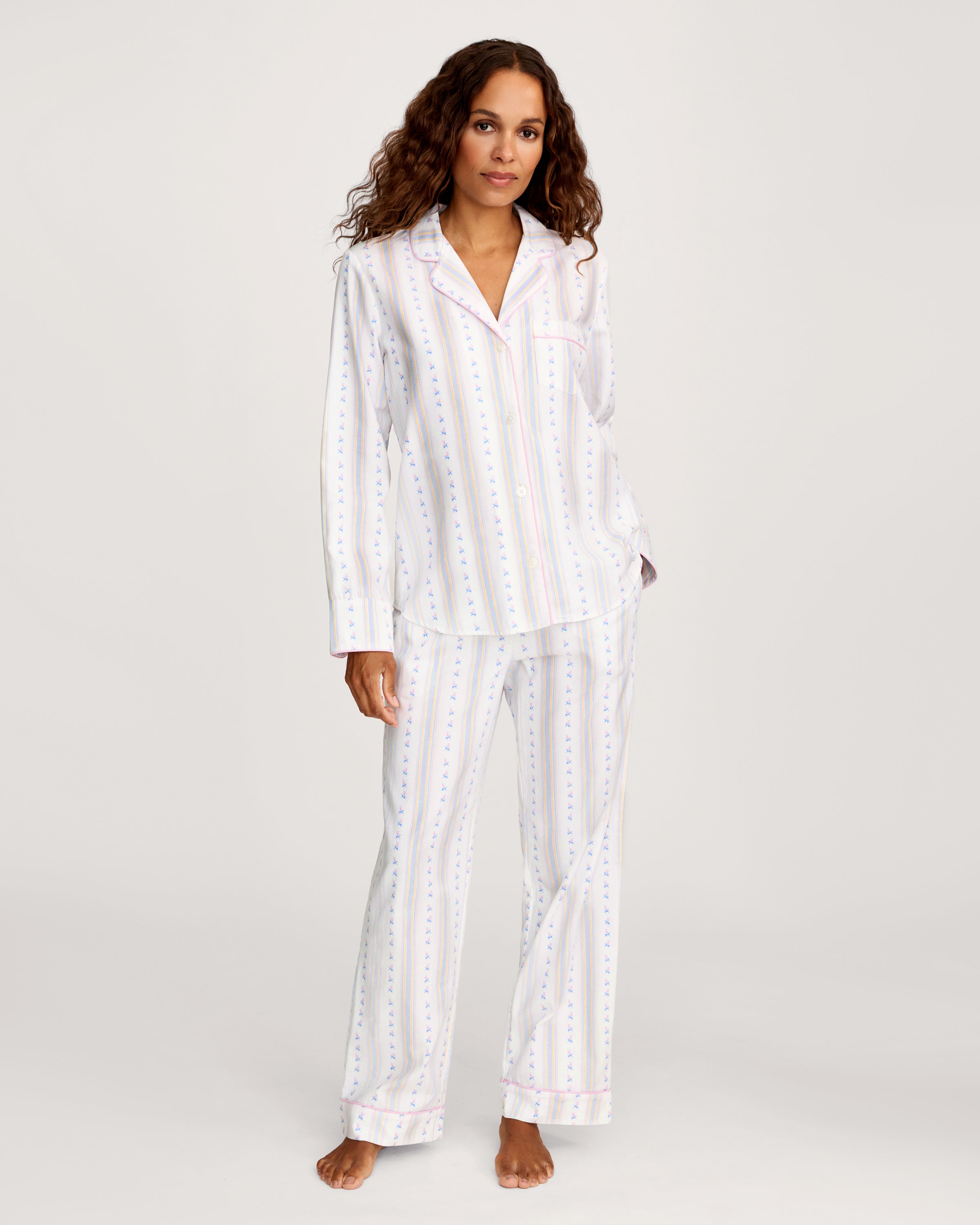 A woman with long curly hair stands barefoot against a plain light background, wearing the Women's Twill Pajama Set in Cottage Stripe—white pajama pants and a matching long-sleeve top with pastel vertical stripes and small dots.