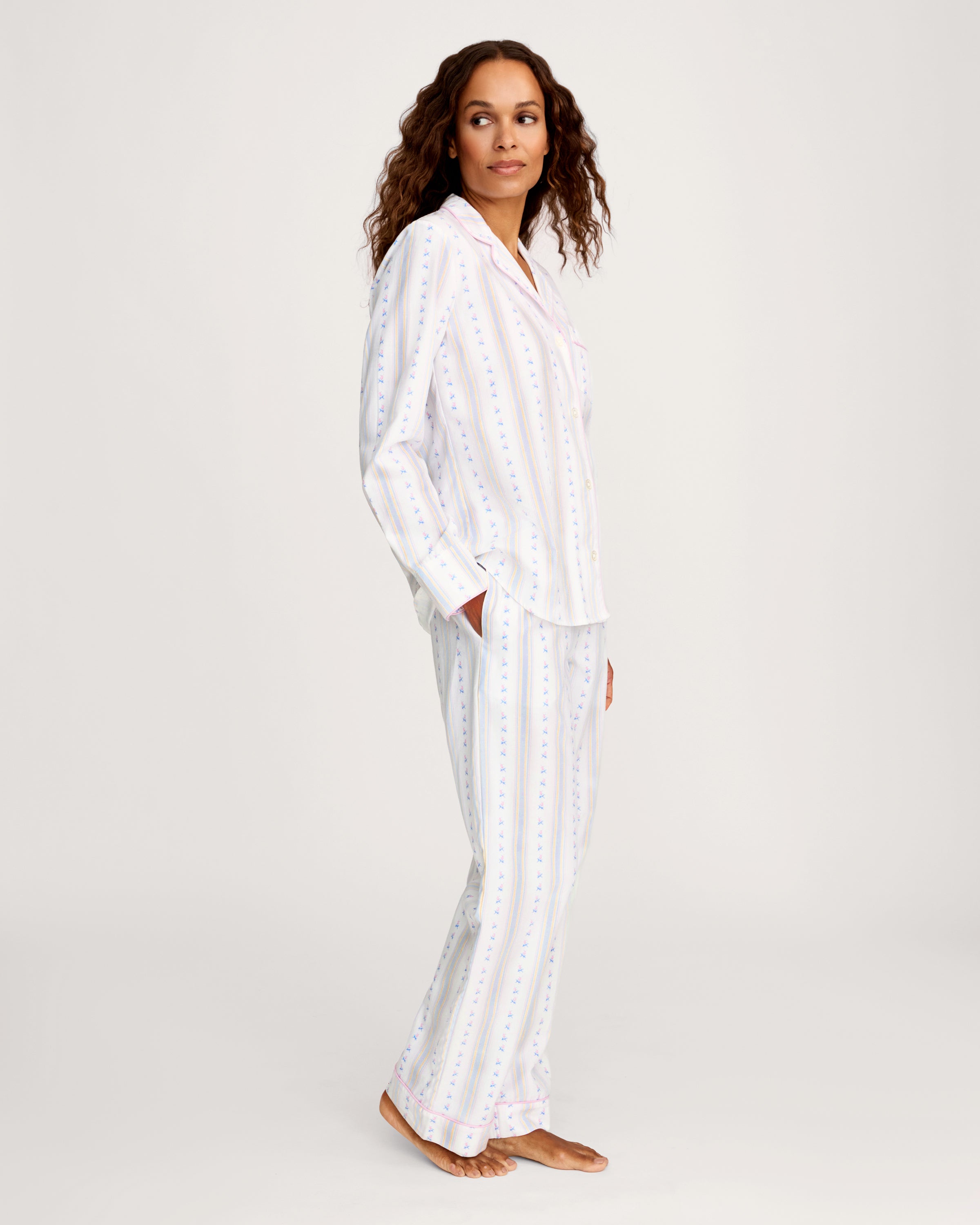 A woman with long curly hair stands barefoot against a plain light background, wearing the Women's Twill Pajama Set in Cottage Stripe—white pajama pants and a matching long-sleeve top with pastel vertical stripes and small dots.