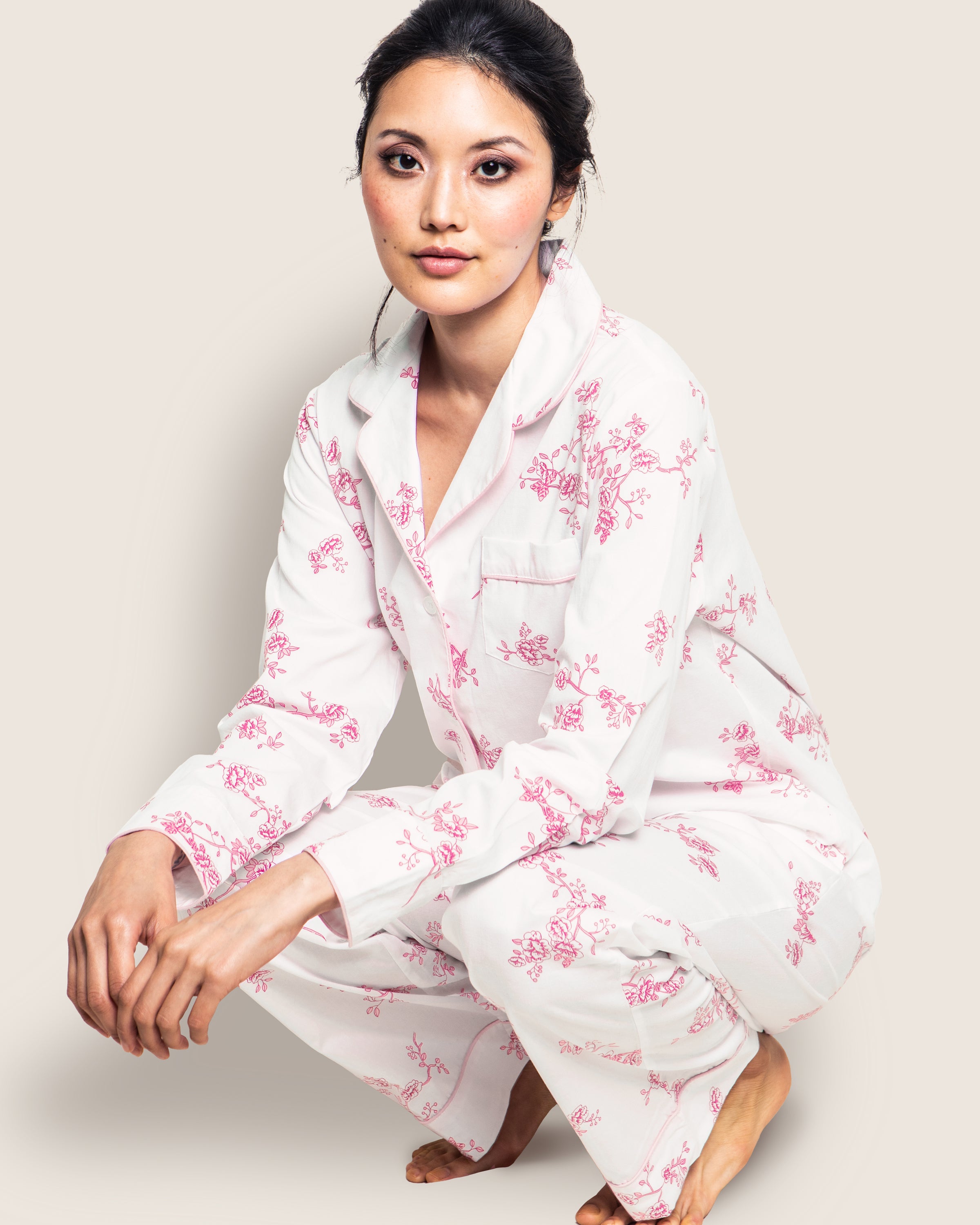 A person crouches in a classic Womens Twill Pajama Set in English Rose Floral by Petite Plume. The finest cotton accentuates a serene expression as they look at the camera against a light-colored backdrop.