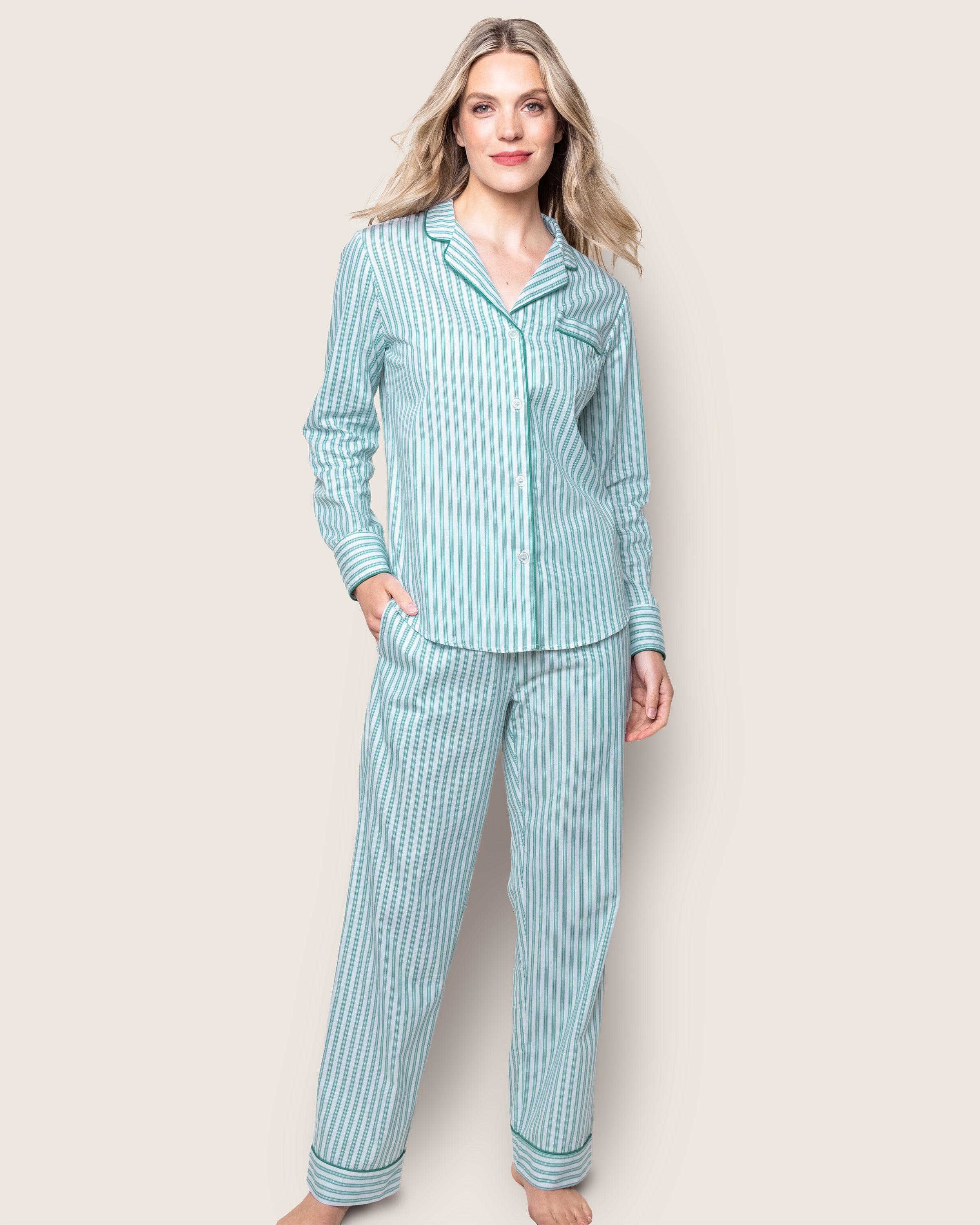 A woman smiles softly, barefoot against a plain background, wearing Petite Plumes Womens Twill Pajama Set in Emerald Ticking. The set features a button-up top and matching pants with a classic French ticking pattern. Her long, wavy hair complements the elegant design.