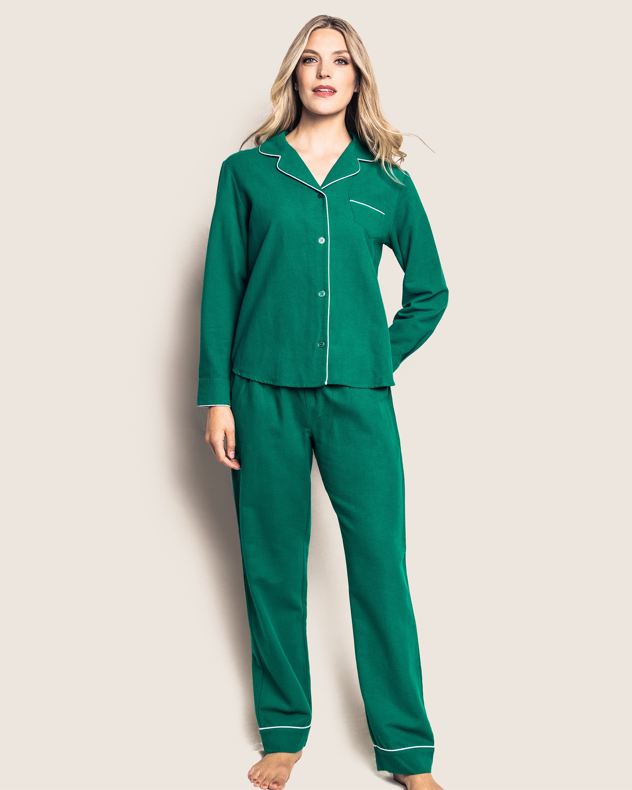 A barefoot person wears the Womens Flannel Pajama Set in Forest Green from Petite Plume, featuring a long-sleeved button-up top with white piping and pockets, paired with matching pants. The background is plain off-white.