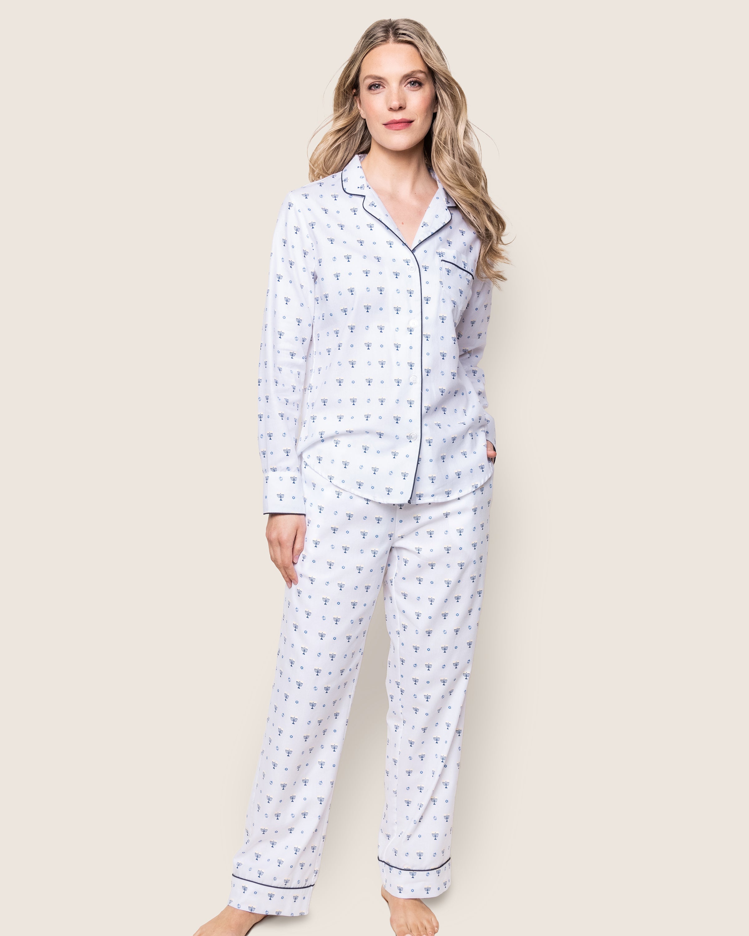 The woman wears Petite Plumes Womens Twill Pajama Set in Happy Hanukkah, featuring a subtle blue menorah design on white, with a collared button-up top and matching pants made of soft cotton twill. Her long, wavy blonde hair cascades as she stands barefoot against a plain light background.