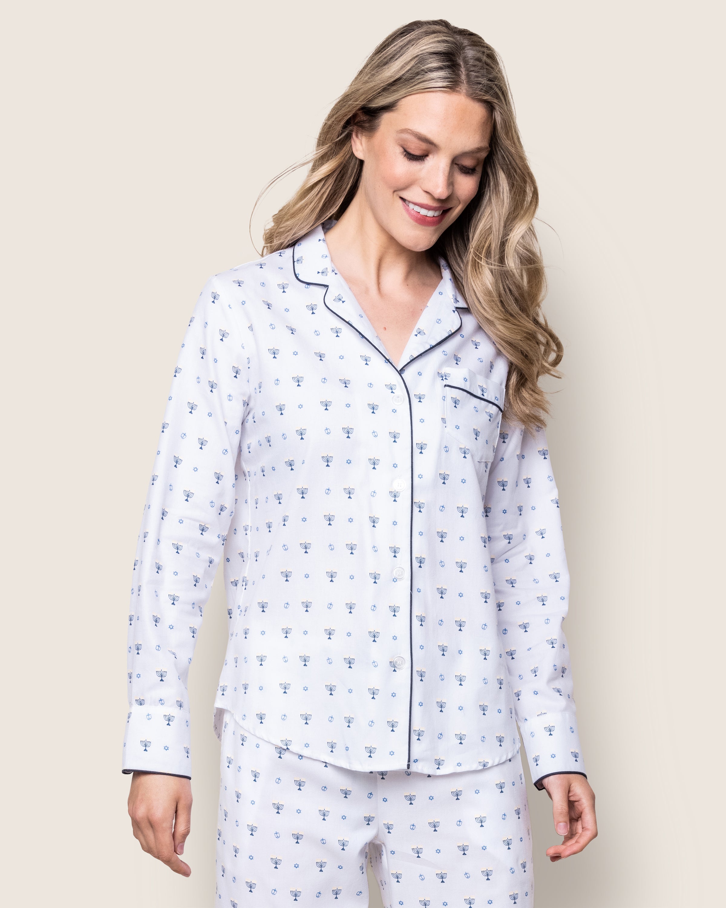 A smiling woman with long blonde hair wears Petite Plumes Womens Twill Pajama Set in Happy Hanukkah, showcasing a subtle menorah design and dark piping. She stands against a plain light background, looking downward with one hand by her side.