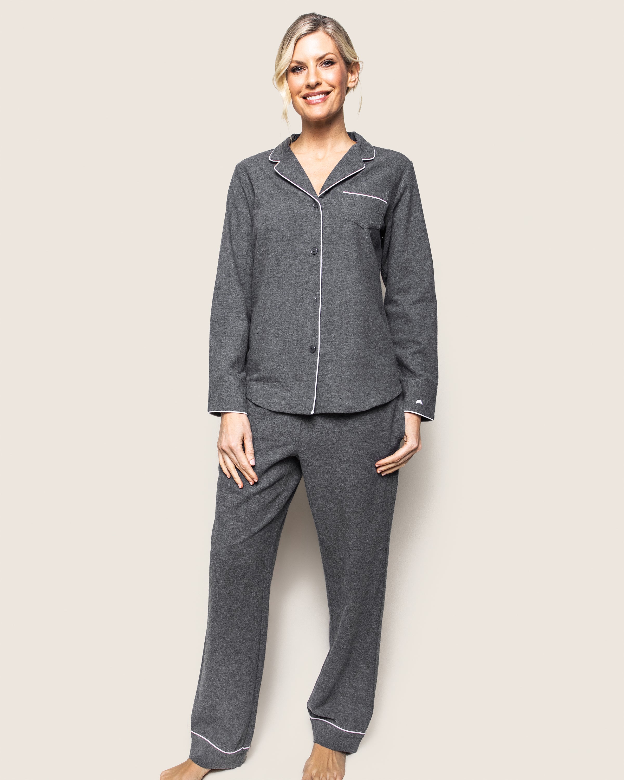 A person wearing the Womens Flannel Pajama Set in Grey by Petite Plume stands barefoot against a plain background. The pajamas, known for their moisture-wicking properties, complement the persons smile and short, light-colored hair.