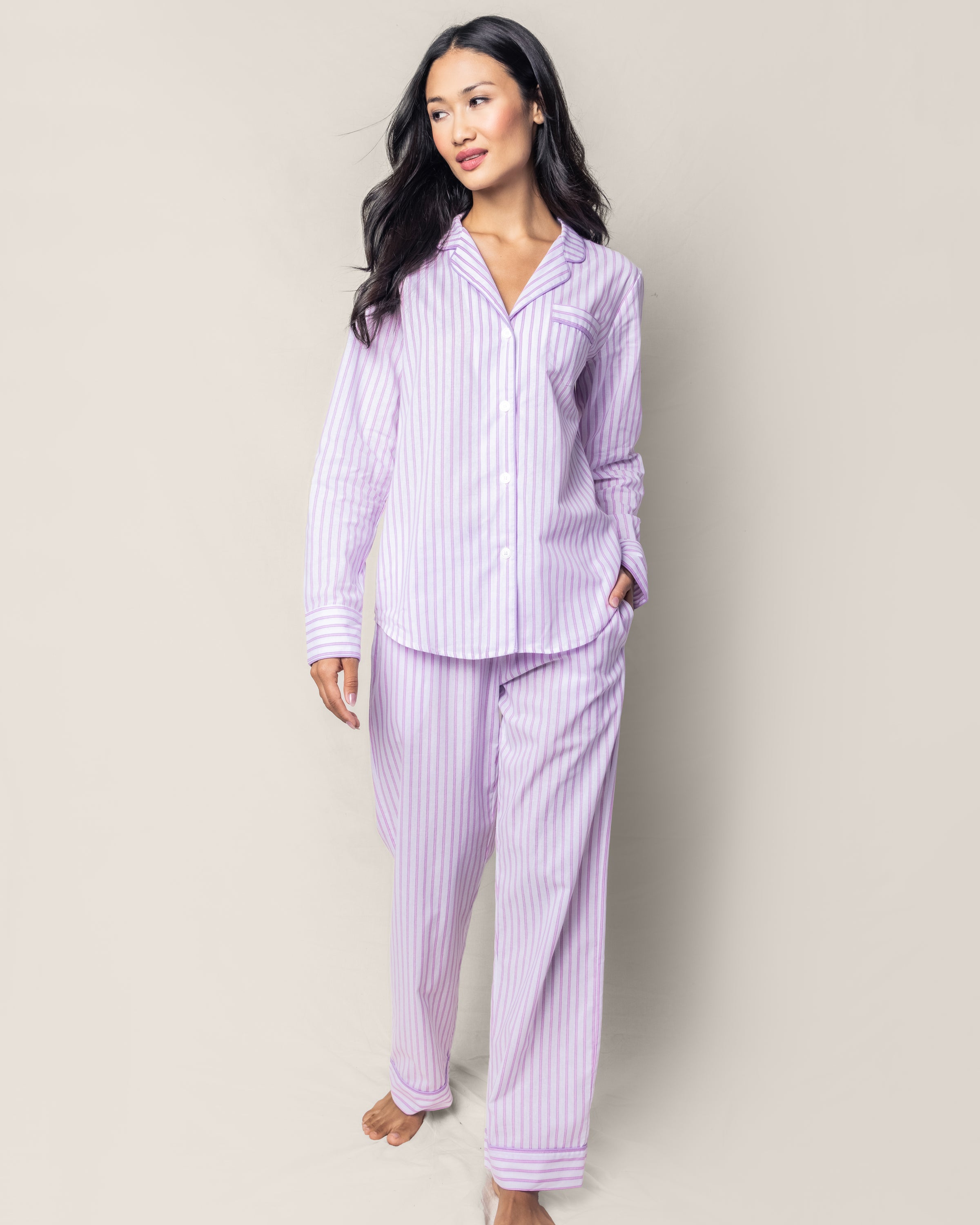 A woman, with long dark hair, dons Petite Plumes Womens Twill Pajama Set in Lavender French Ticking. The high-quality cotton top features a collar and buttons as she tucks her hands into her pants pockets against a plain light gray background.