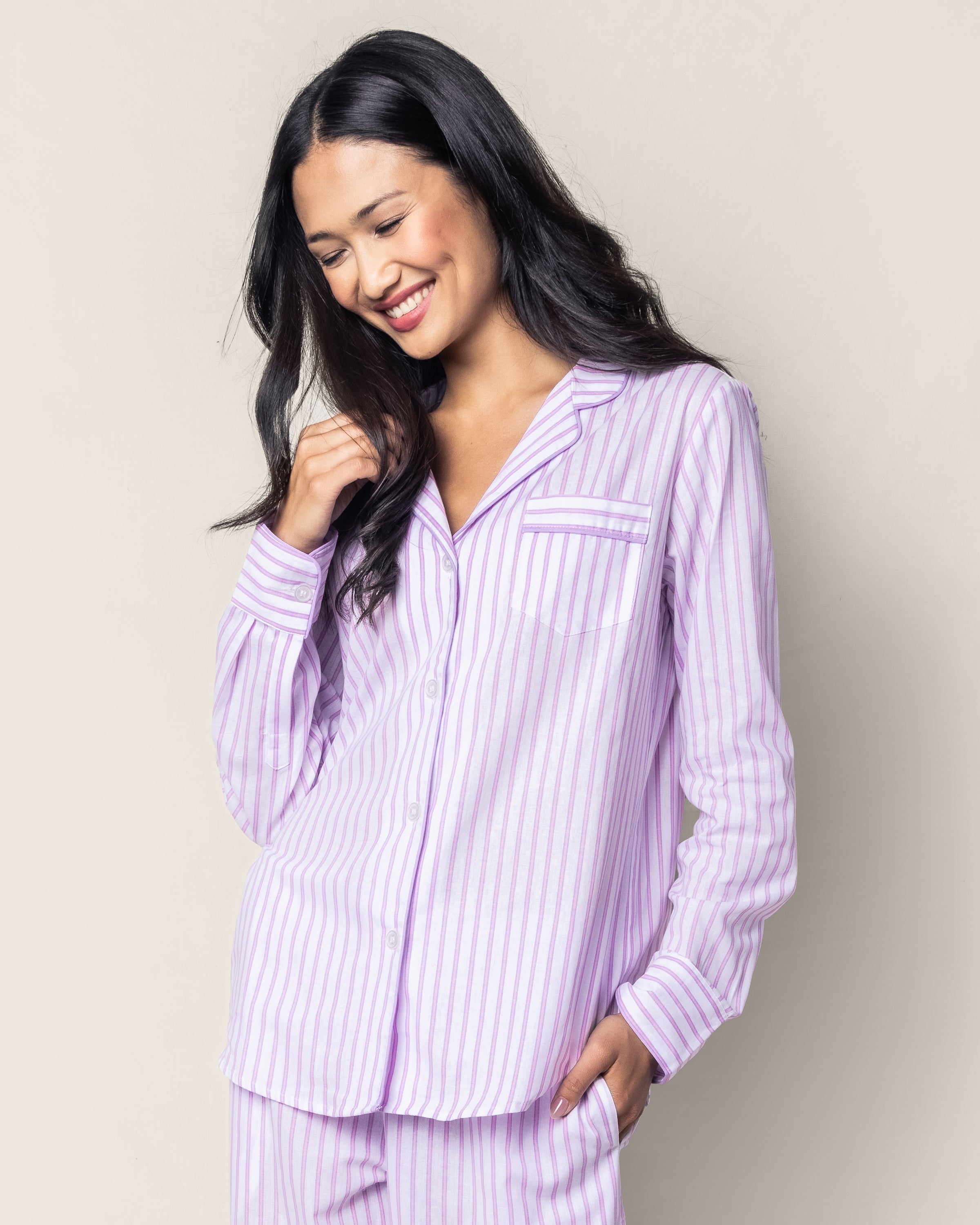 A person with long dark hair smiles in a Petite Plume Womens Twill Pajama Set in Lavender French Ticking, against a plain, light background, looking down while holding their hair.