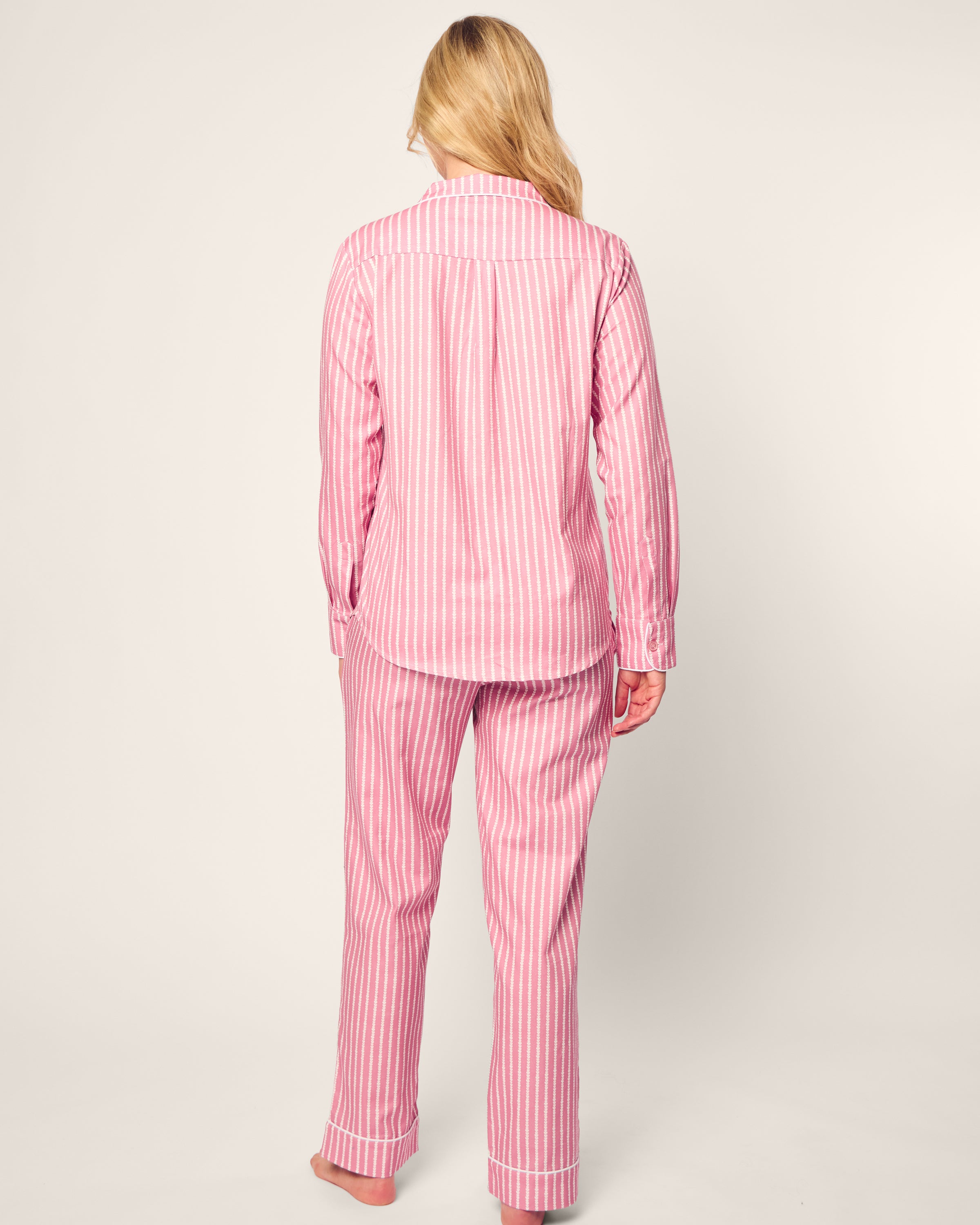 A person with long blonde hair is seen from behind in pink and white striped pajamas from Petite Plumes Love Lines collection. Made from high-quality cotton, the Womens Twill Pajama Set includes a long-sleeved top and pants, set against a simple light-colored background.