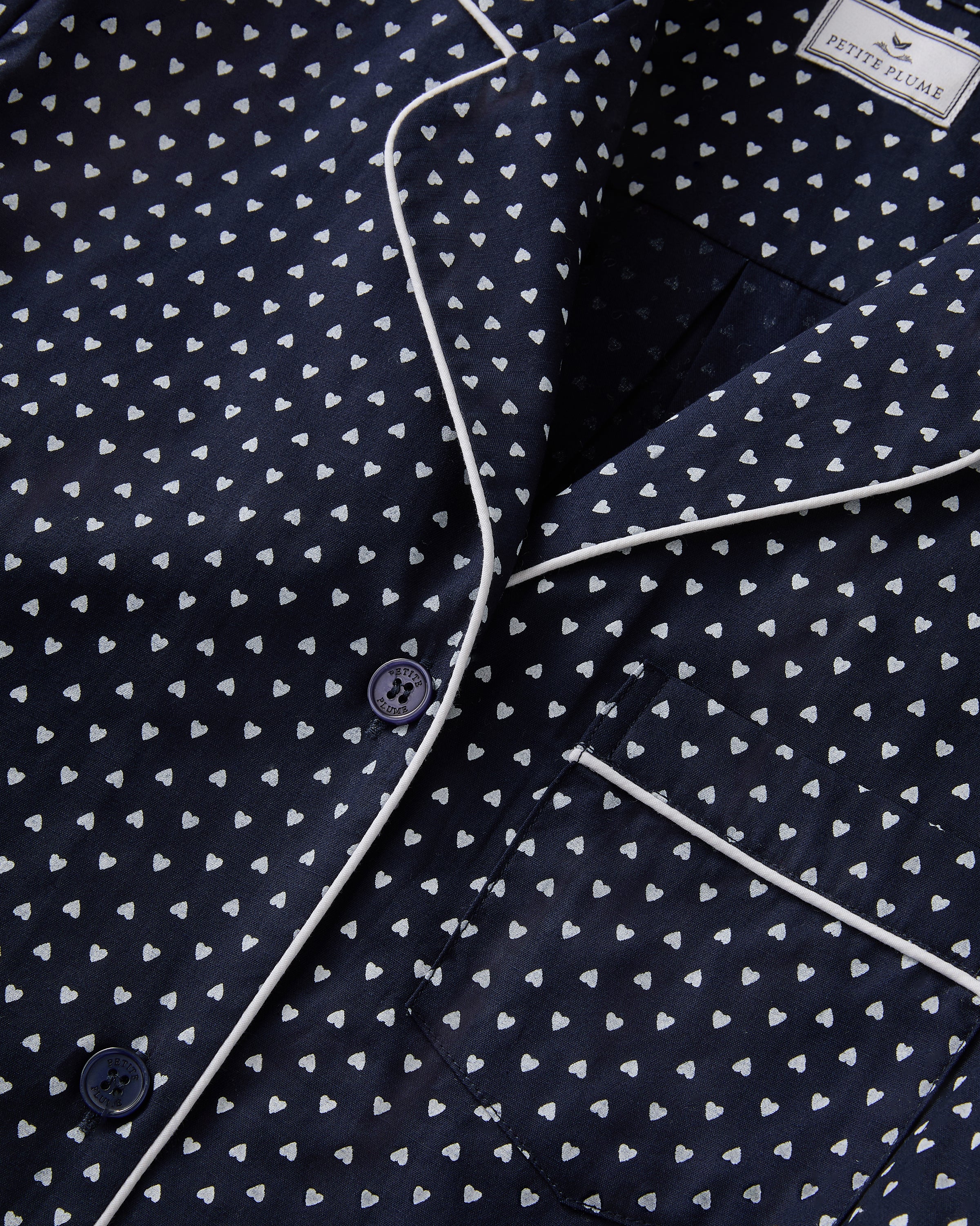 The Women's Twill Pajama Set in Midnight Hearts features a navy blue, long-sleeve top with a collar, button-down front, white heart print, and white piping on the edges and pocket, paired with matching pants.