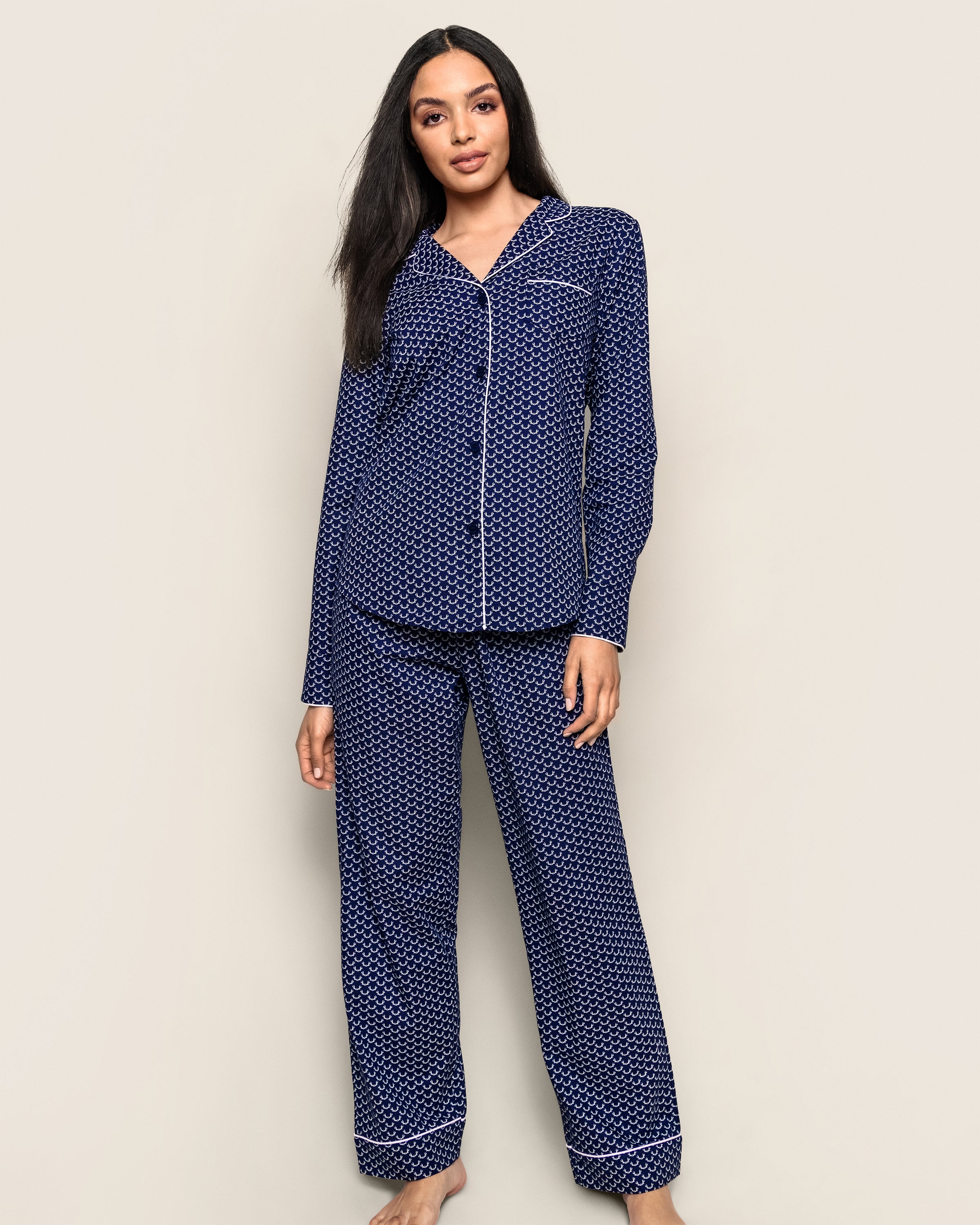 A woman stands barefoot against a light background, wearing Petite Plumes Womens Twill Pajama Set in navy Nordic Antlers, featuring elegant white piping.