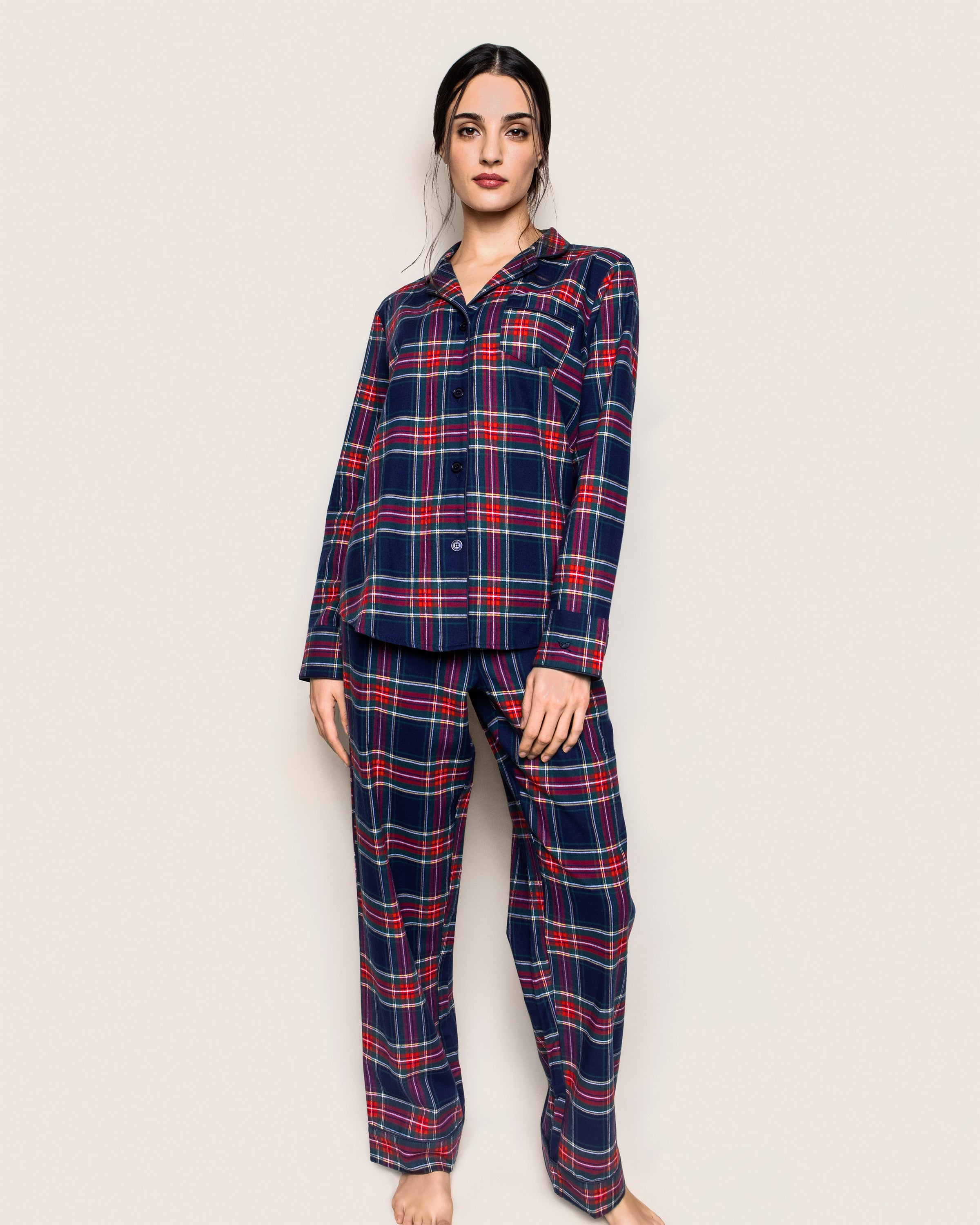 Against a beige background, someone stands barefoot in Petite Plumes Womens Brushed Cotton Pajama Set in Windsor Tartan—crafted from 100% cotton twill, featuring a dark blue base with red and green stripes. The moisture-wicking weave, collared top, and buttons ensure comfort.