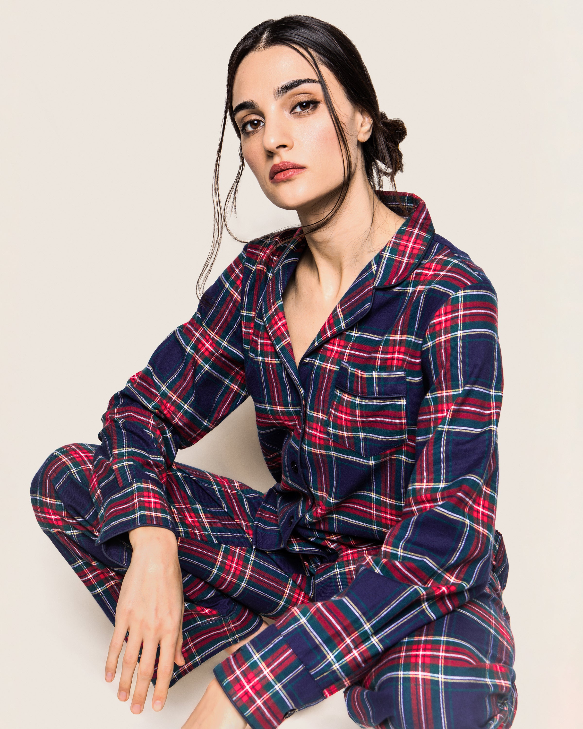 A person in a Womens Brushed Cotton Pajama Set in Windsor Tartan by Petite Plume sits with one knee up against a plain background, hair styled in a low bun with loose strands framing the face, and a neutral expression.