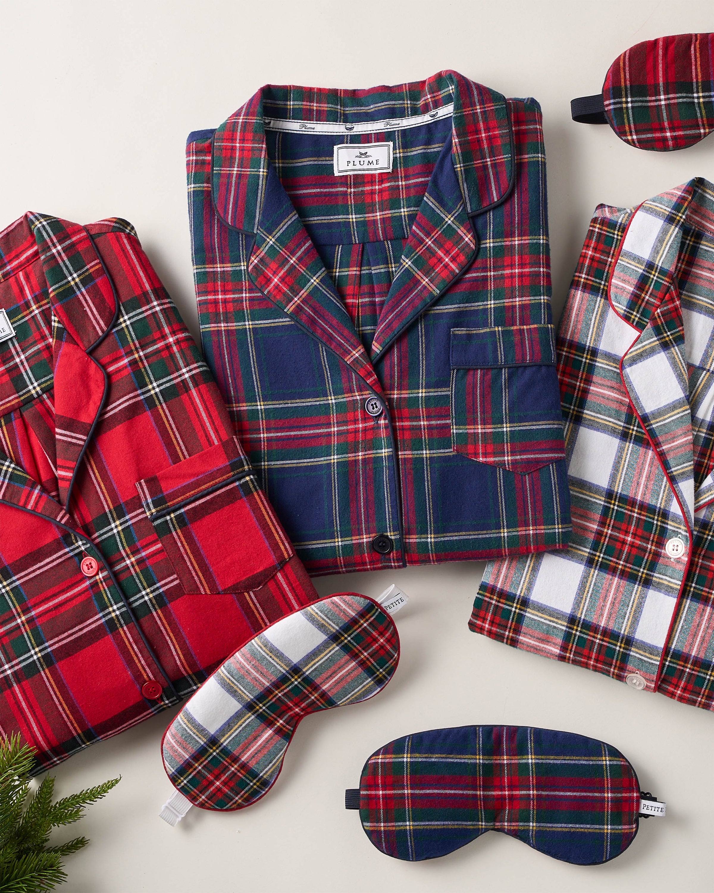 Petite Plumes Womens Brushed Cotton Pajama Set in Windsor Tartan, featuring button-up tops with front pockets, is displayed alongside matching sleep masks on a light surface. Made from 100% cotton twill for comfort and style, a small green branch decorates the corner.