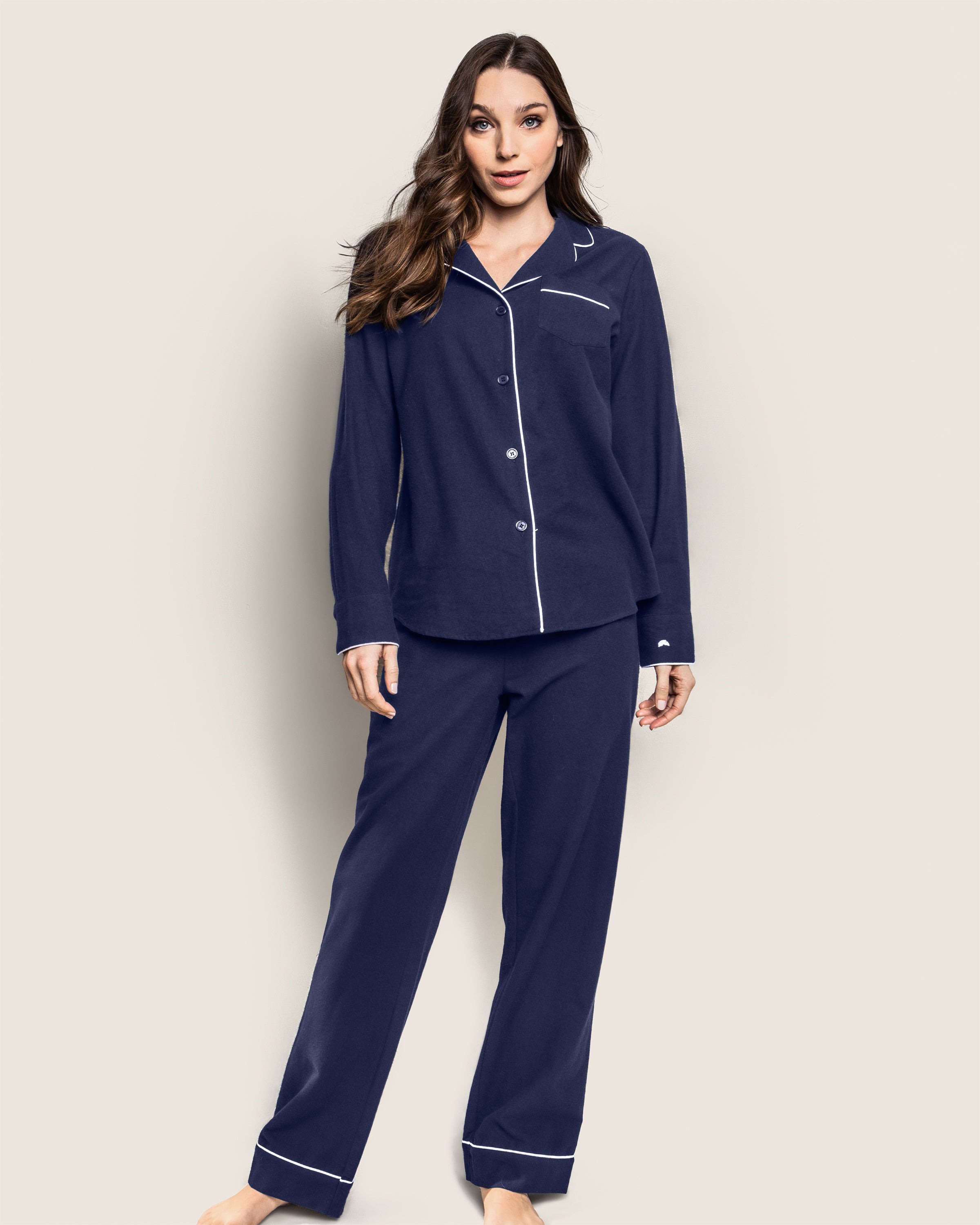 A woman in Petite Plumes Womens Flannel Pajama Set in Navy with white piping stands barefoot against a light background. The cozy ensemble includes a button-up top and matching pants, embodying effortless relaxation with her long hair cascading down.
