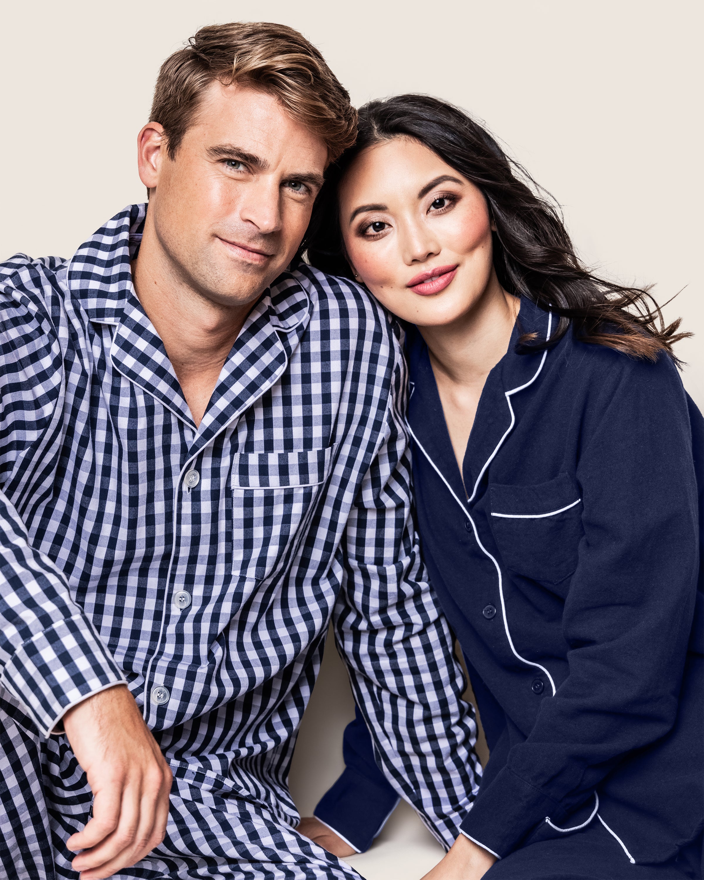 A man in blue and white checkered pajamas stands beside a woman in a Womens Flannel Pajama Set in Navy by Petite Plume. She rests her head softly on his shoulder, both displaying serene expressions against a neutral background.