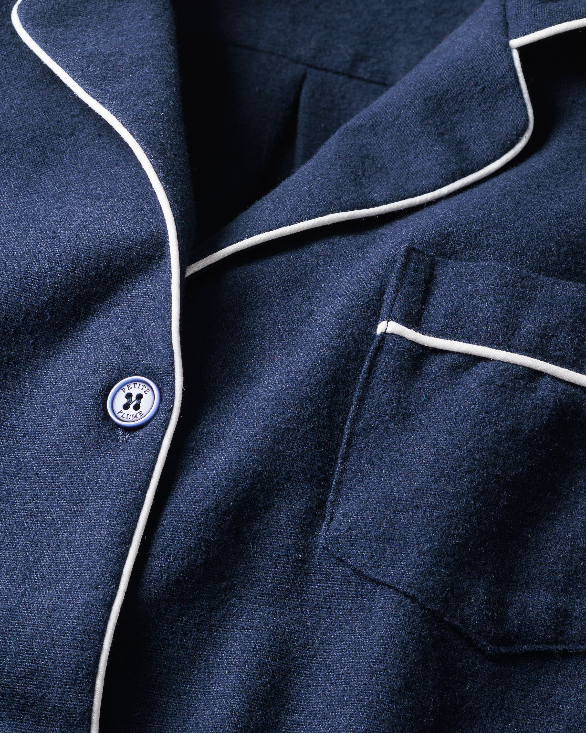 The Womens Flannel Pajama Set in Navy by Petite Plume is shown in a close-up with white piping on the collar and pocket edges. A button with a paw print design is near the collar, highlighting the soft and smooth fabric.