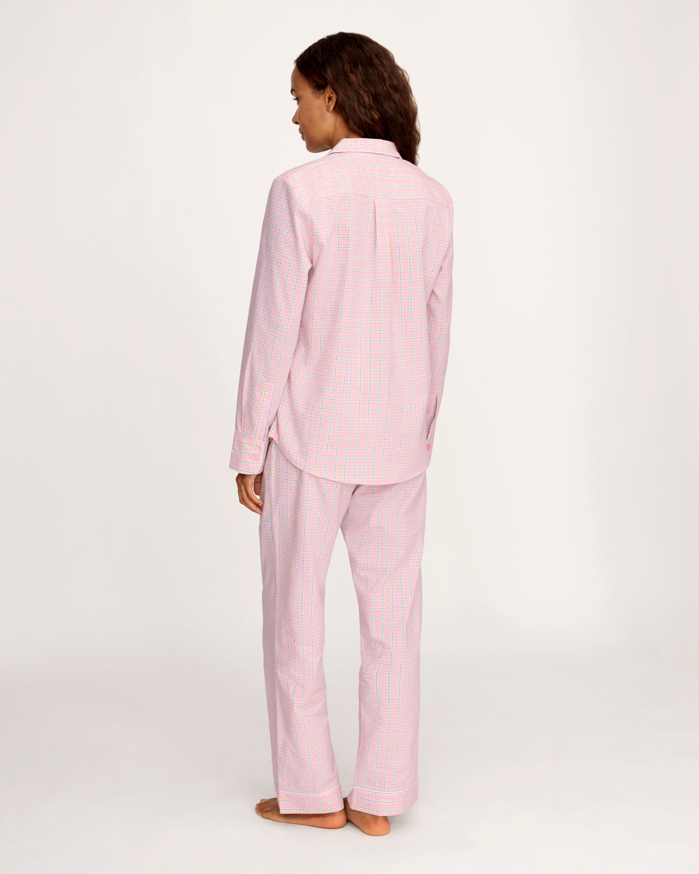 A woman with long curly hair wears the Women's Twill Pajama Set in Peony Plaid, standing barefoot against a plain light background and facing the camera with a relaxed expression.