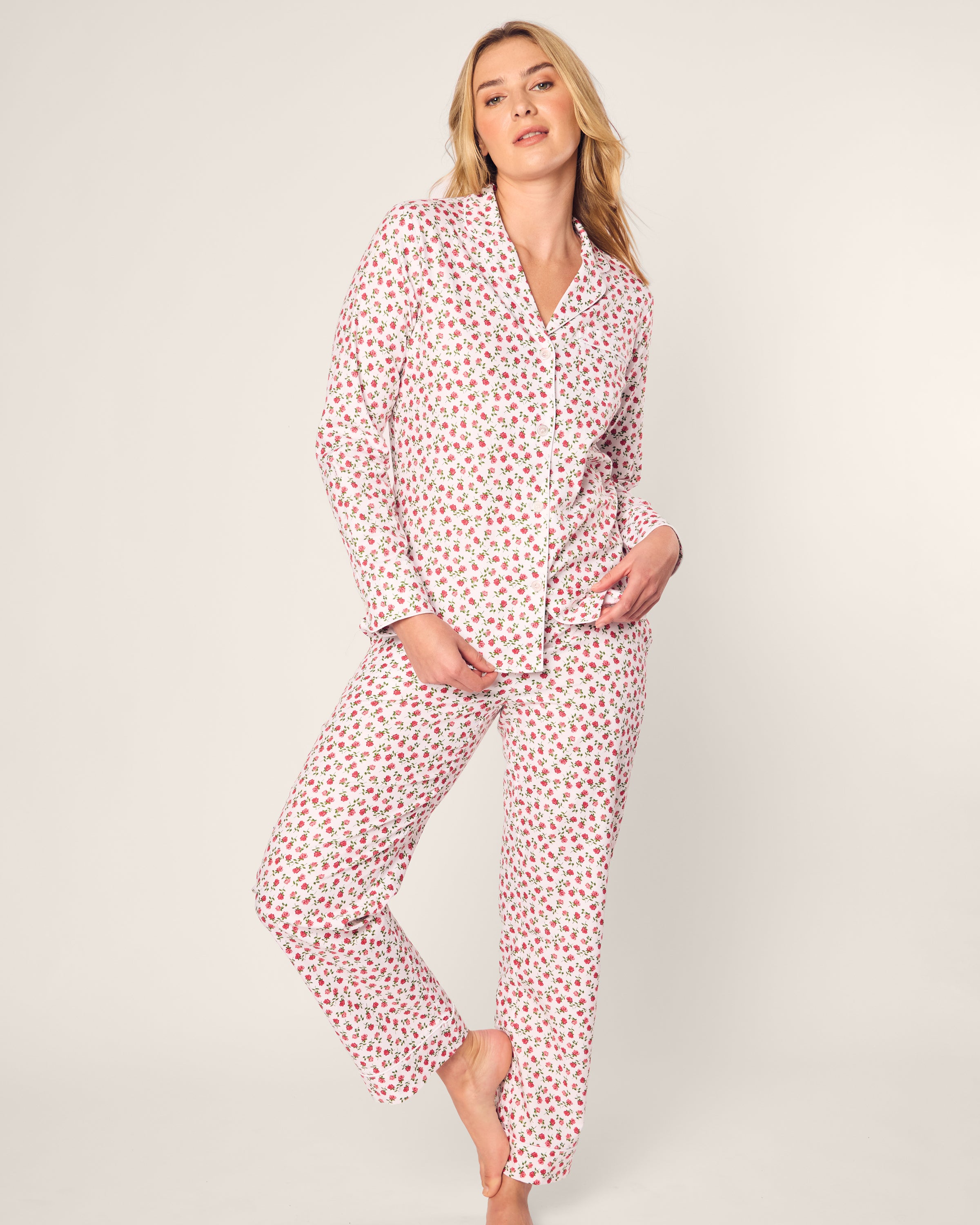 A person in luxurious sleepwear poses against a plain background, wearing Petite Plumes Womens Twill Pajama Set in Petite Petals, made from high-quality cotton. The design features white fabric with small pink flowers, and the person stands relaxed, one hand in a pocket and the other on their thigh.